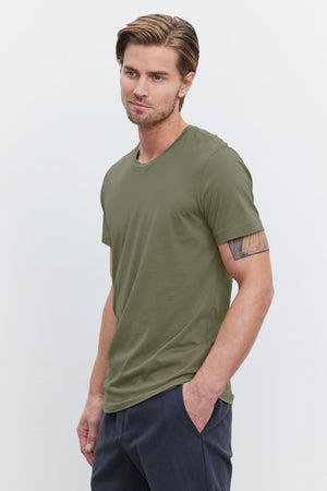 A young man with light brown hair wearing an olive green HOWARD TEE from Velvet by Graham & Spencer made from lightweight cotton knit and dark pants stands with his hands in his pockets against a plain white background, giving off a perfect fit.