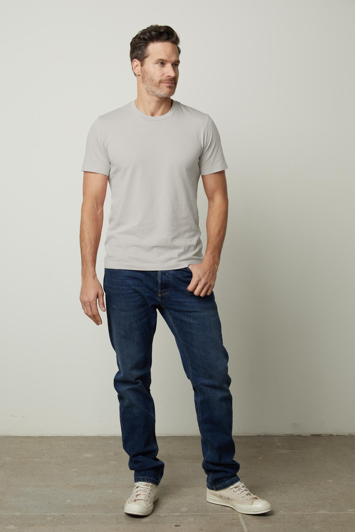   A man stands against a plain background, wearing a light gray HOWARD TEE by Velvet by Graham & Spencer, blue jeans, and white sneakers. The shirt's vintage feel adds character while offering the perfect fit. 