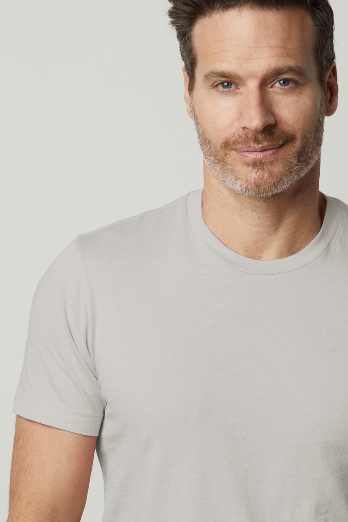   Man with short dark hair and light stubble, wearing the HOWARD TEE by Velvet by Graham & Spencer, made of Pima cotton jersey in a plain light gray color, smiling slightly in front of a neutral background. 