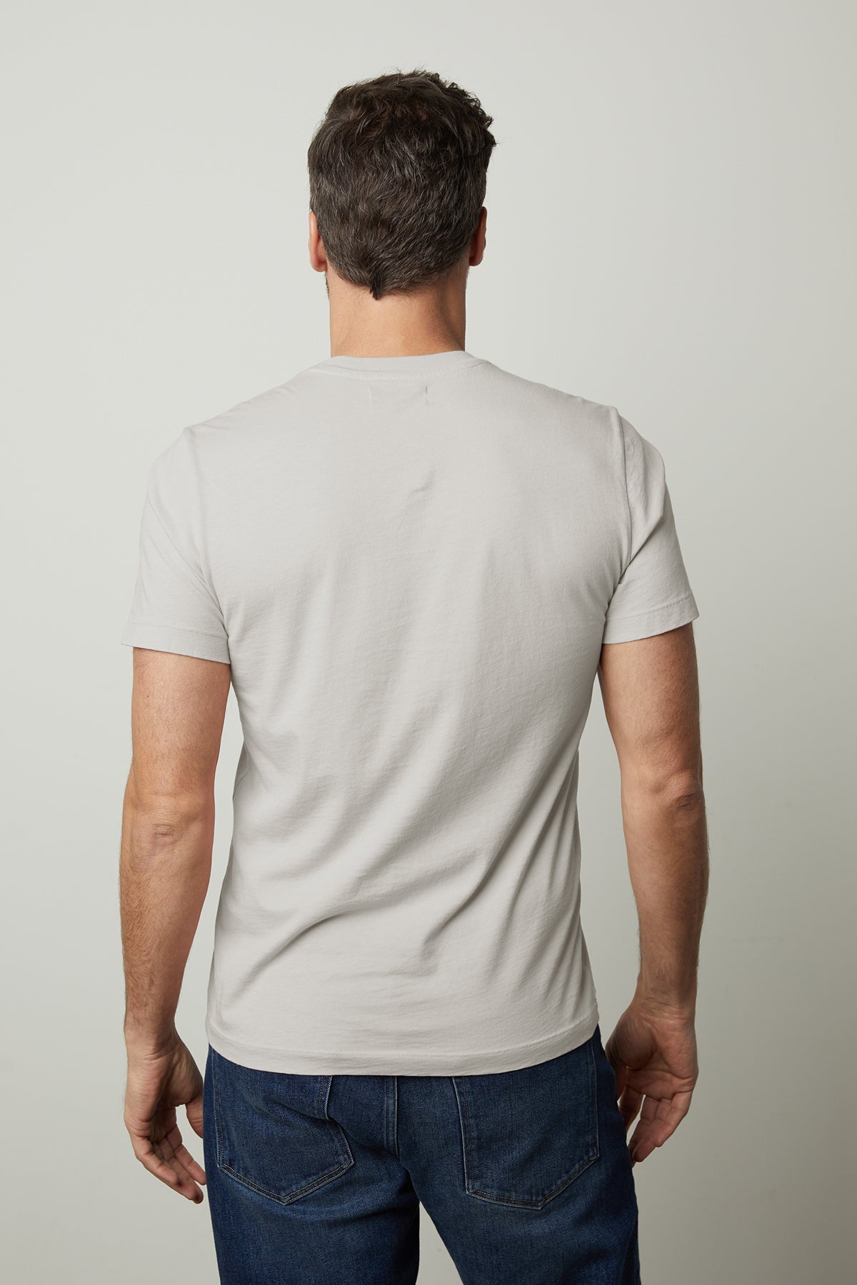 A man with short, dark hair stands with his back to the camera, wearing a light gray Velvet by Graham & Spencer HOWARD TEE made from lightweight cotton knit and dark blue jeans, offering a perfect fit.-37229772734657