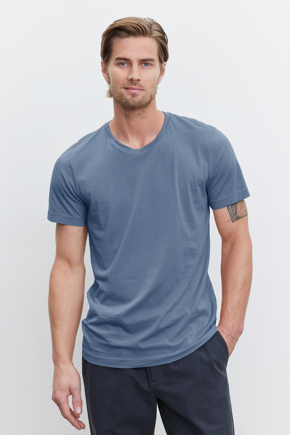   A man with short, light brown hair and a beard is wearing the HOWARD TEE made of Pima cotton jersey by Velvet by Graham & Spencer in plain blue and dark pants, standing against a white background. 