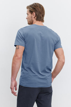 A man with light brown hair is standing with his back to the camera, wearing a perfectly fitted HOWARD TEE from Velvet by Graham & Spencer, made of soft Pima cotton jersey in a striking blue color, paired with dark pants for a vintage feel.