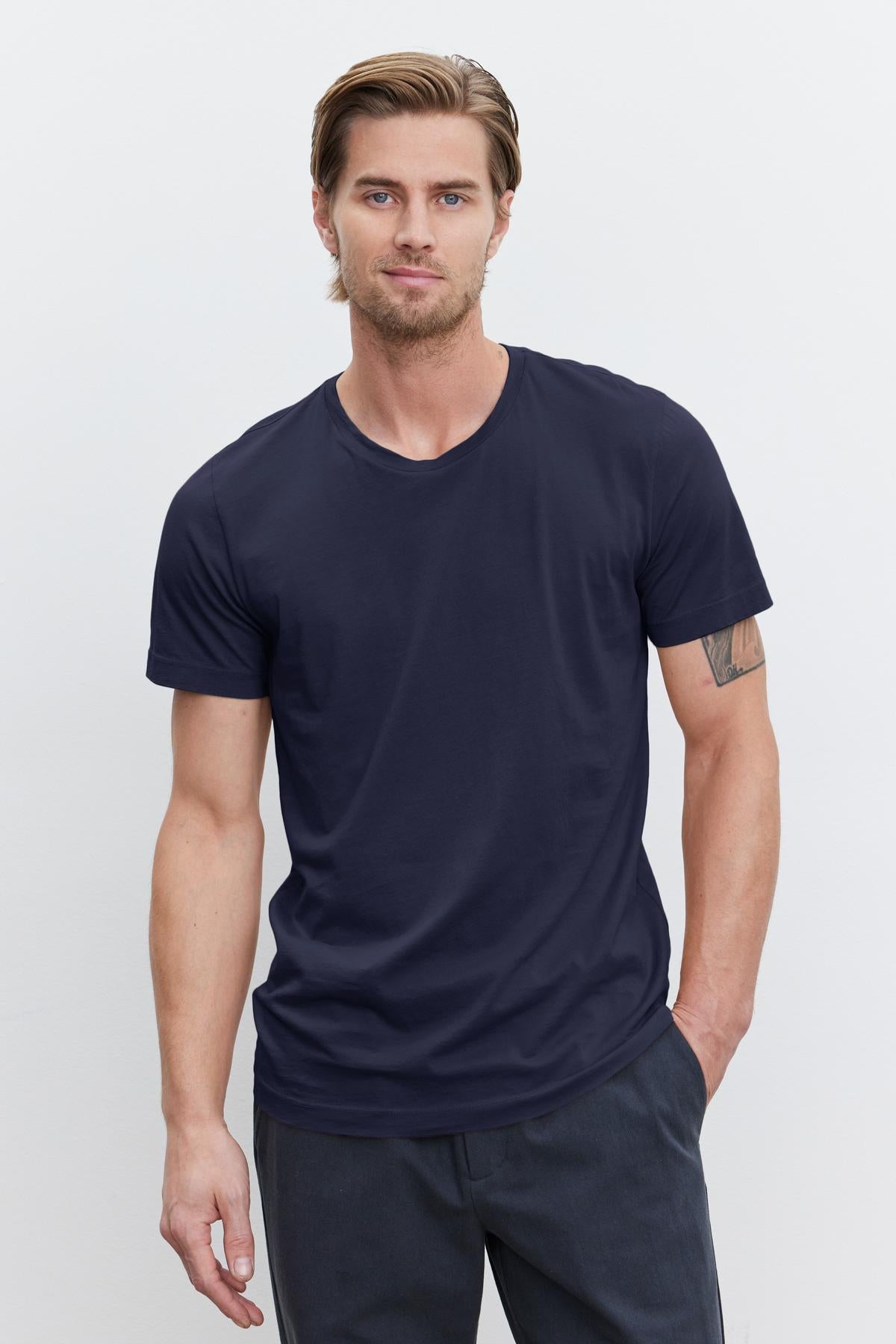   A man with light brown hair and a beard is wearing a navy blue HOWARD TEE from Velvet by Graham & Spencer made from lightweight cotton knit and dark pants, standing against a plain white background. He has one hand in his pocket. 