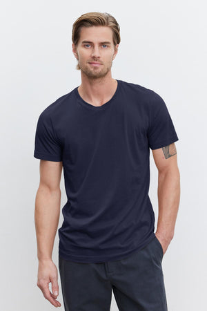 A man with light brown hair and a beard is wearing a navy blue HOWARD TEE from Velvet by Graham & Spencer made from lightweight cotton knit and dark pants, standing against a plain white background. He has one hand in his pocket.