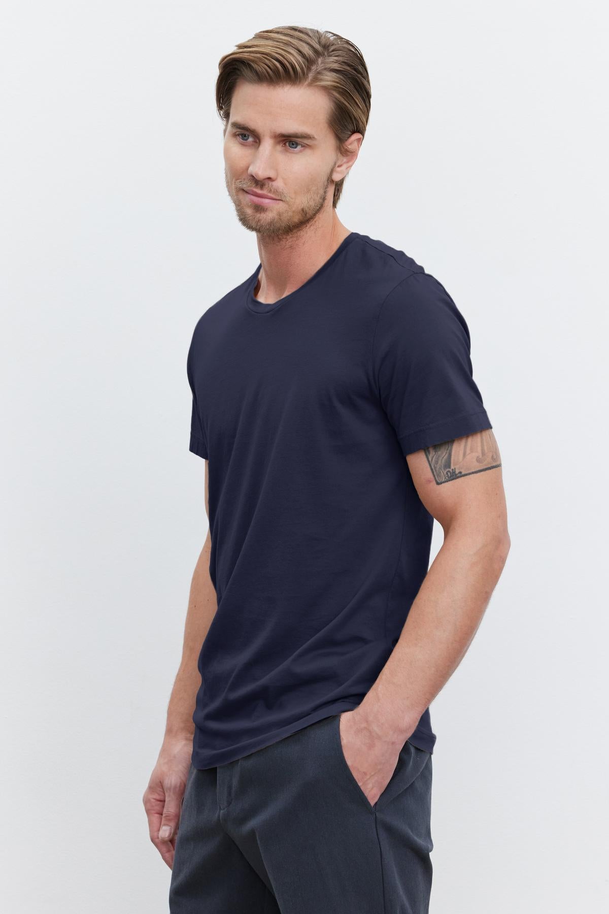   Man standing against a plain white background, wearing a navy blue lightweight cotton knit HOWARD TEE by Velvet by Graham & Spencer and dark trousers with his left hand in his pocket. He has short brown hair and an arm tattoo. The HOWARD TEE has a vintage feel and offers the perfect fit. 