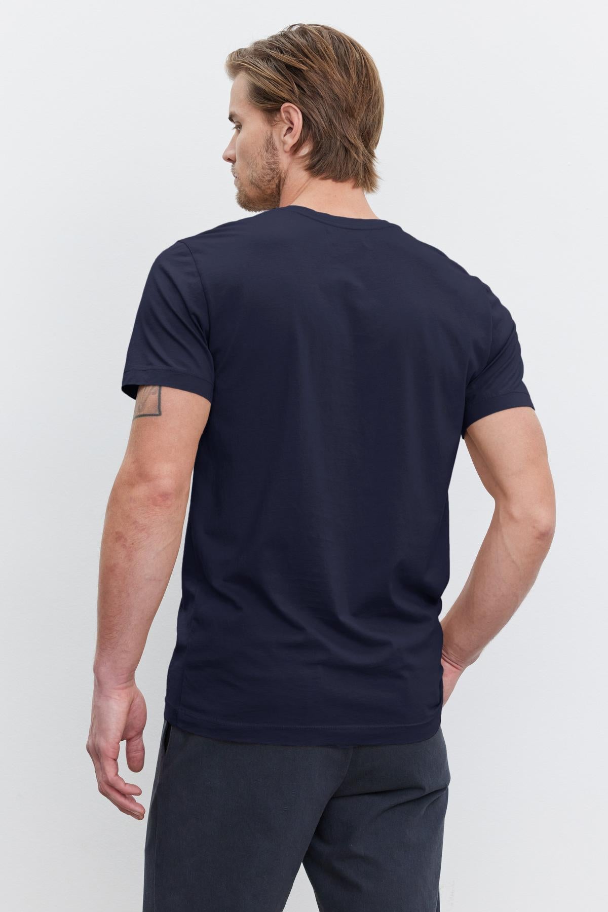 A man with short hair and a beard, wearing a Velvet by Graham & Spencer HOWARD TEE and gray pants, stands with his back to the camera. His right arm features a tattoo.-36273879384257