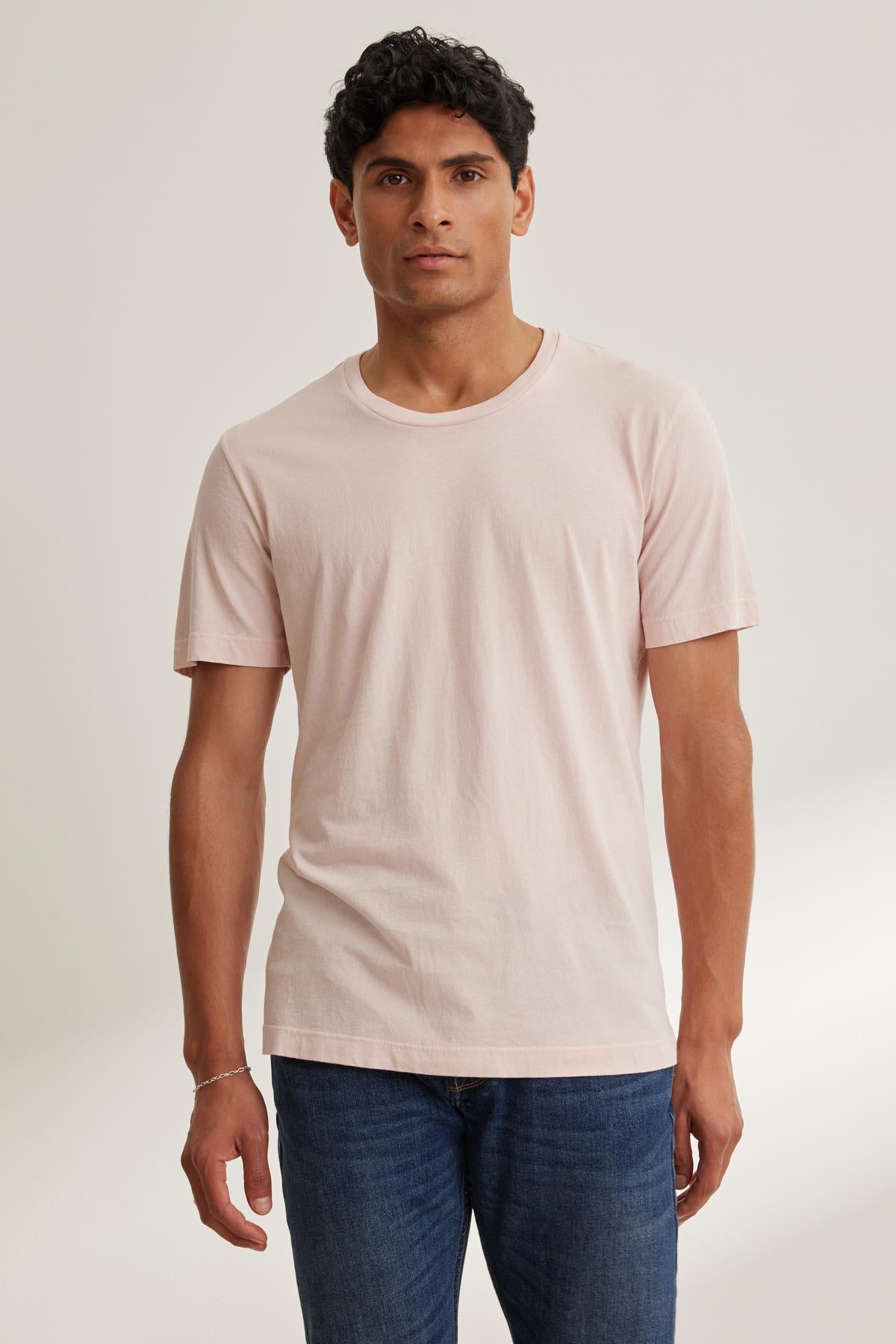   Against a neutral background, a person wears the Velvet by Graham & Spencer HOWARD TEE in light pink, crafted from lightweight Pima jersey, paired with blue jeans for a perfect vintage fit. 