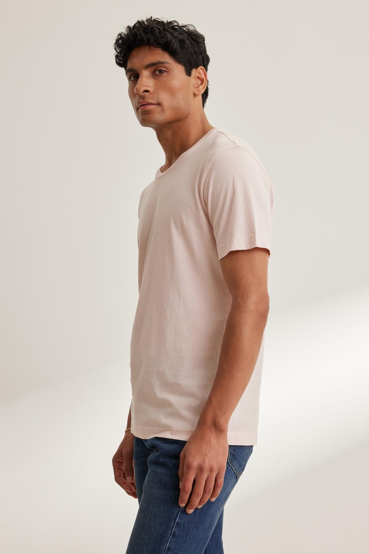   A man stands sideways in a Velvet by Graham & Spencer HOWARD TEE, made from lightweight Pima jersey, paired with blue jeans. His outfit's perfect fit and the plain background create a timeless vintage vibe as he gazes ahead. 