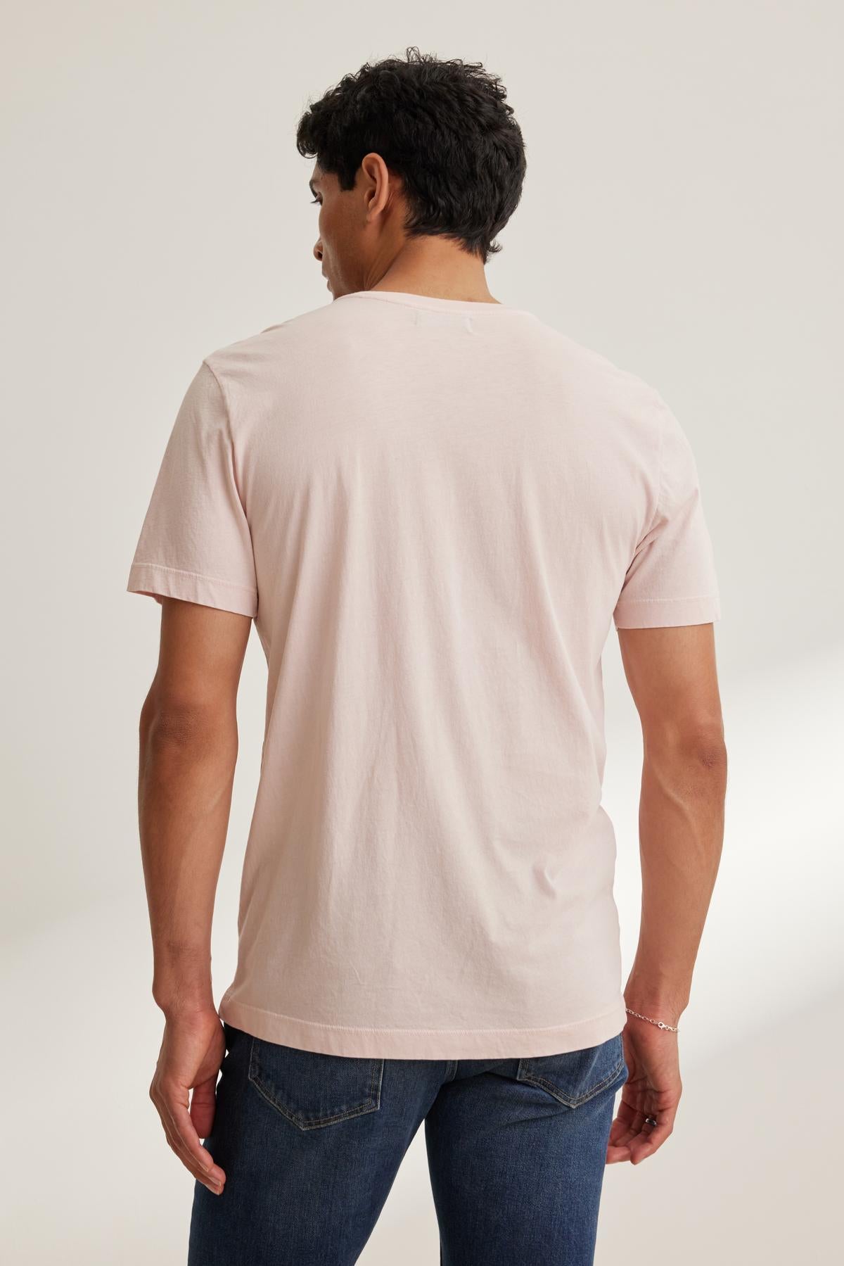   In a well-lit room, a person wearing the HOWARD TEE in light pink by Velvet by Graham & Spencer, paired with blue jeans, stands facing away. 