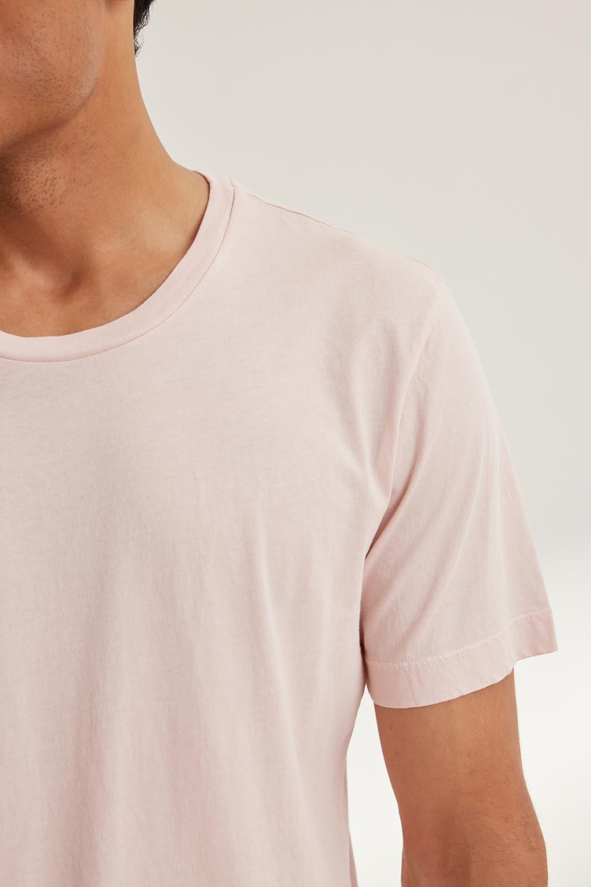   A person wearing the HOWARD TEE by Velvet by Graham & Spencer, featuring a vintage light pink Pima jersey design, stands against a neutral background. The lightweight fabric offers a perfect fit, complementing their style effortlessly. 