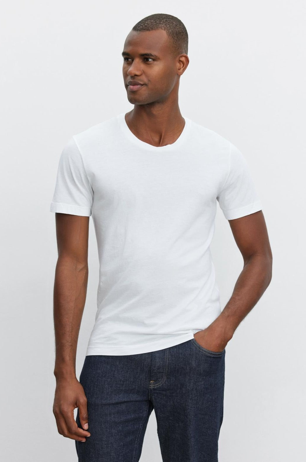   A man stands against a plain background, wearing a white HOWARD TEE made by Velvet by Graham & Spencer and dark blue jeans. He is looking slightly to the side and has one hand in his pocket, exuding a vintage feel. 