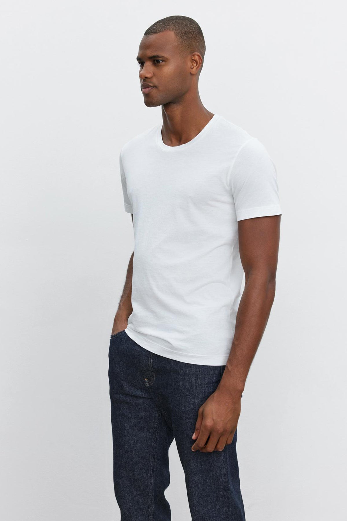   A man stands against a plain, light-colored background, wearing a white HOWARD TEE by Velvet by Graham & Spencer that boasts a perfect fit and dark jeans, with his left hand in his pocket. 