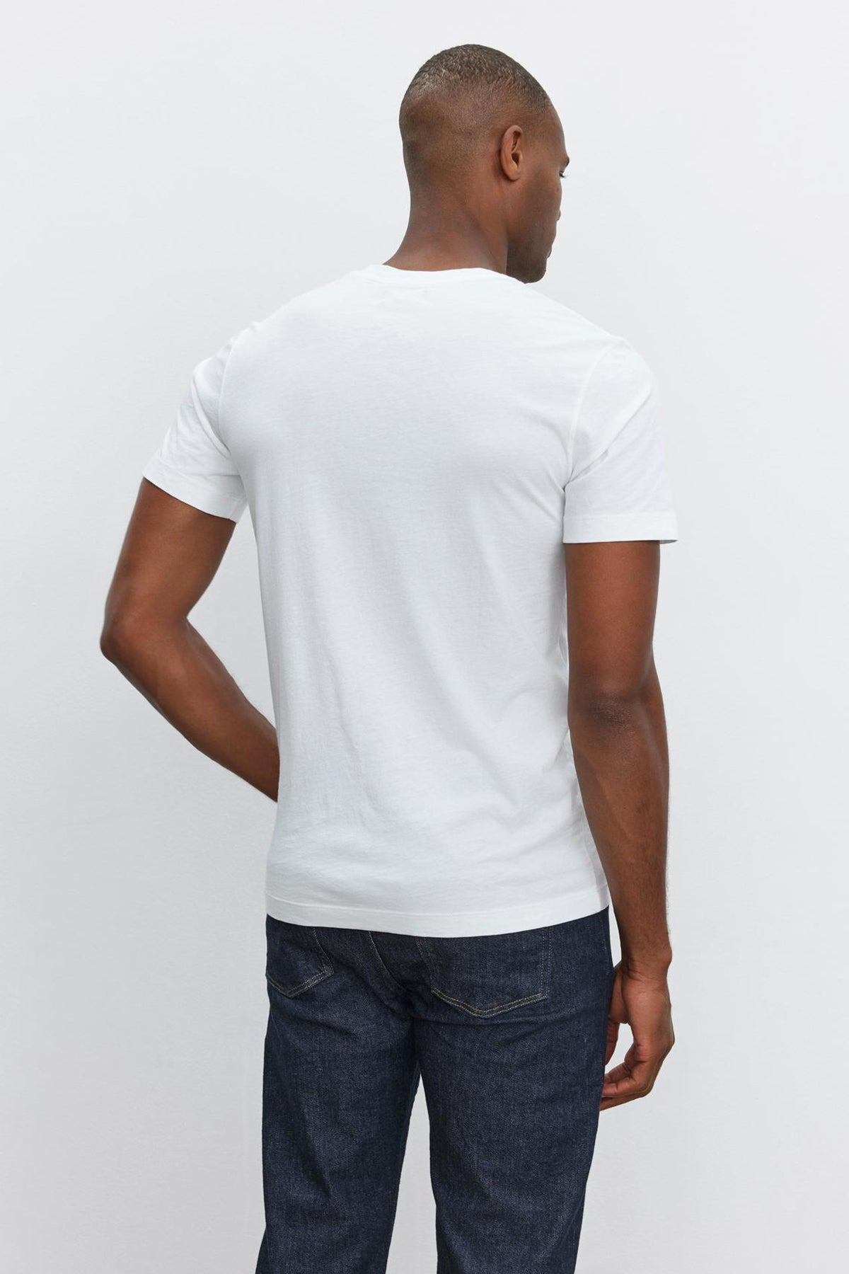   A person is standing with their back to the camera, wearing a plain white HOWARD TEE made from lightweight cotton knit by Velvet by Graham & Spencer and dark blue jeans. 