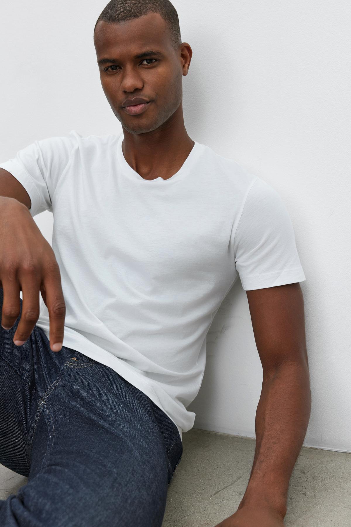   A man in a white HOWARD TEE by Velvet by Graham & Spencer and blue jeans, made from lightweight cotton knit for that perfect fit, sits against a white wall, resting one arm on his knee. 