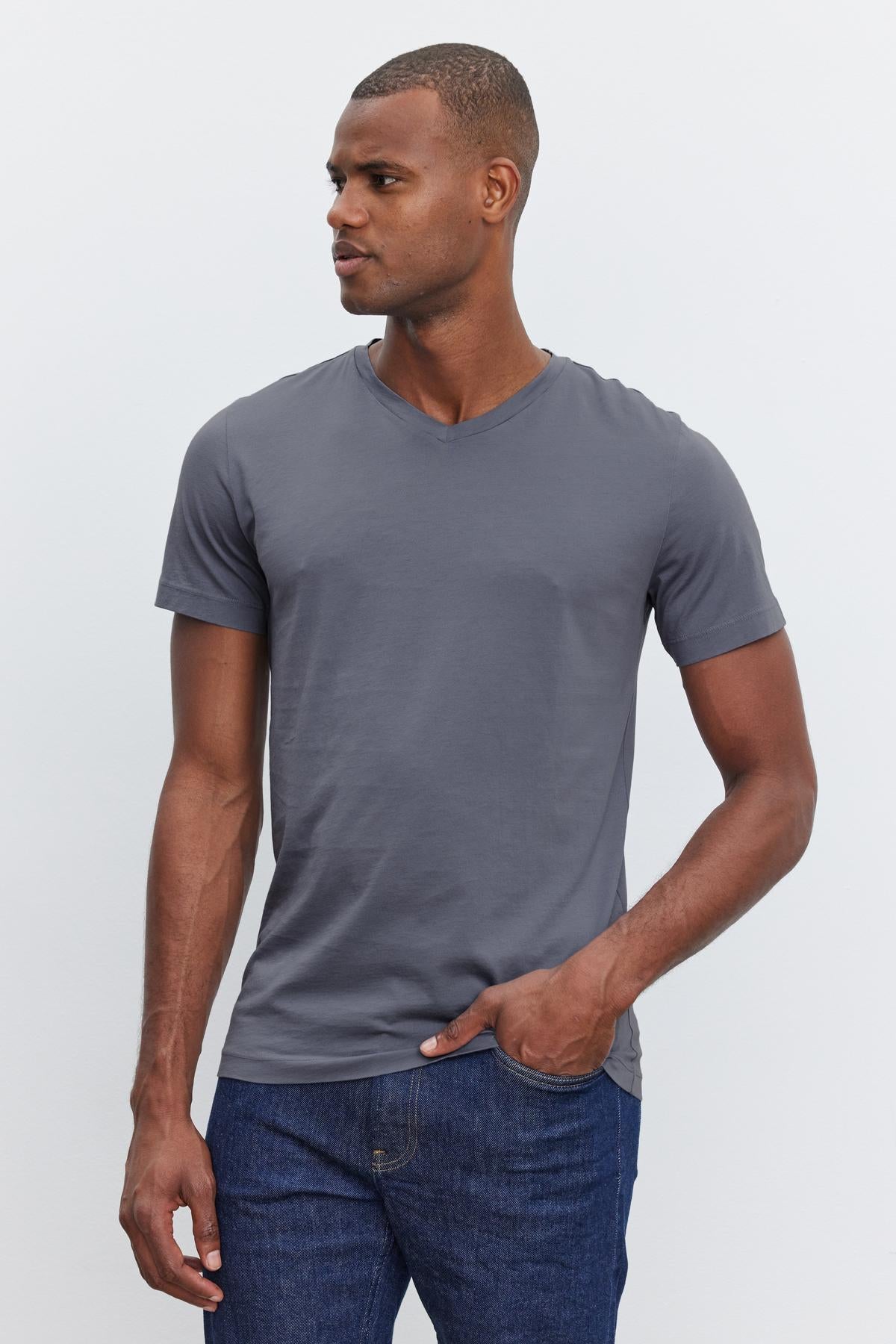   A person in a SAMSEN TEE by Velvet by Graham & Spencer with a v-neckline and blue jeans stands with one hand in their pocket, looking to the side against a plain white background—perfect for everyday wear. 