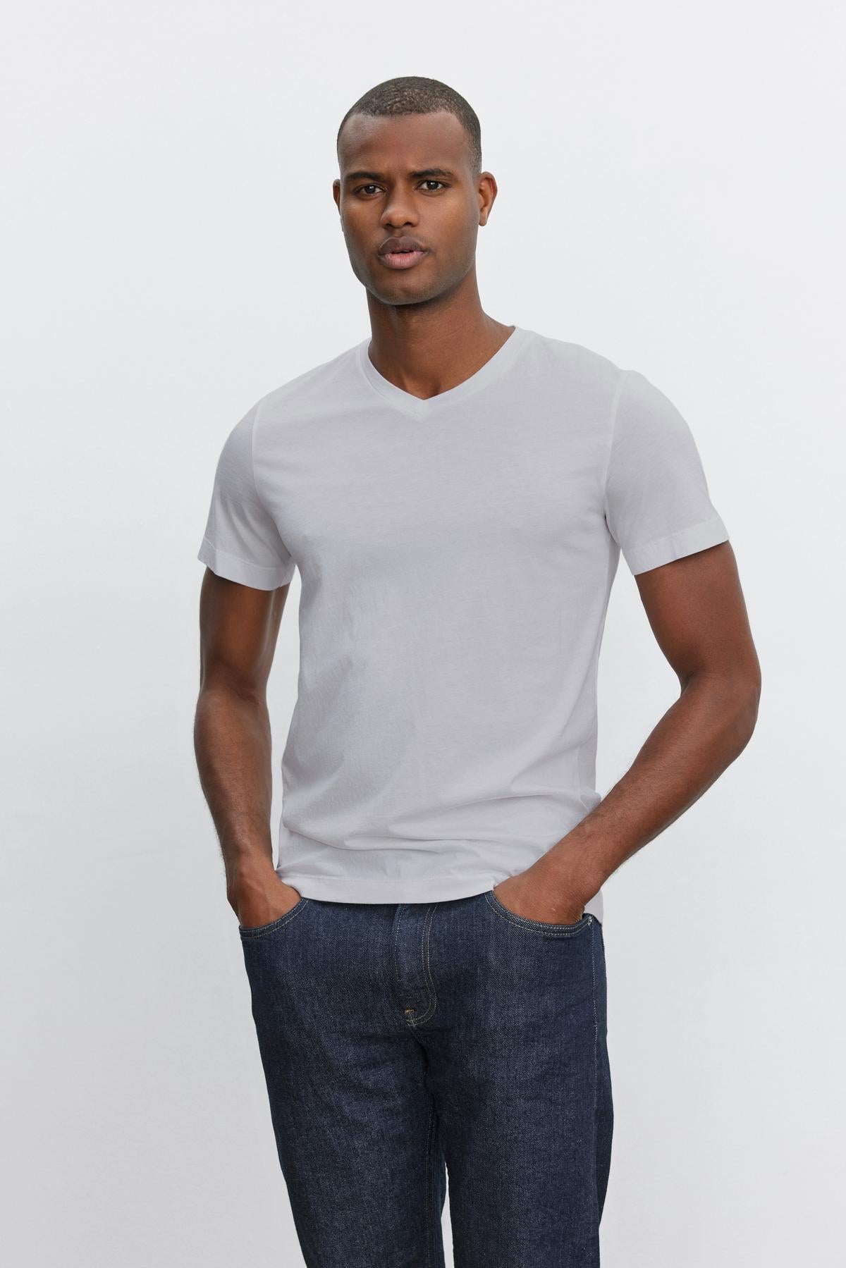   A person is standing with their hands in their pockets, wearing a plain white Velvet by Graham & Spencer SAMSEN TEE with a V-neckline and dark blue jeans—perfect for everyday wear—against a plain white background. 