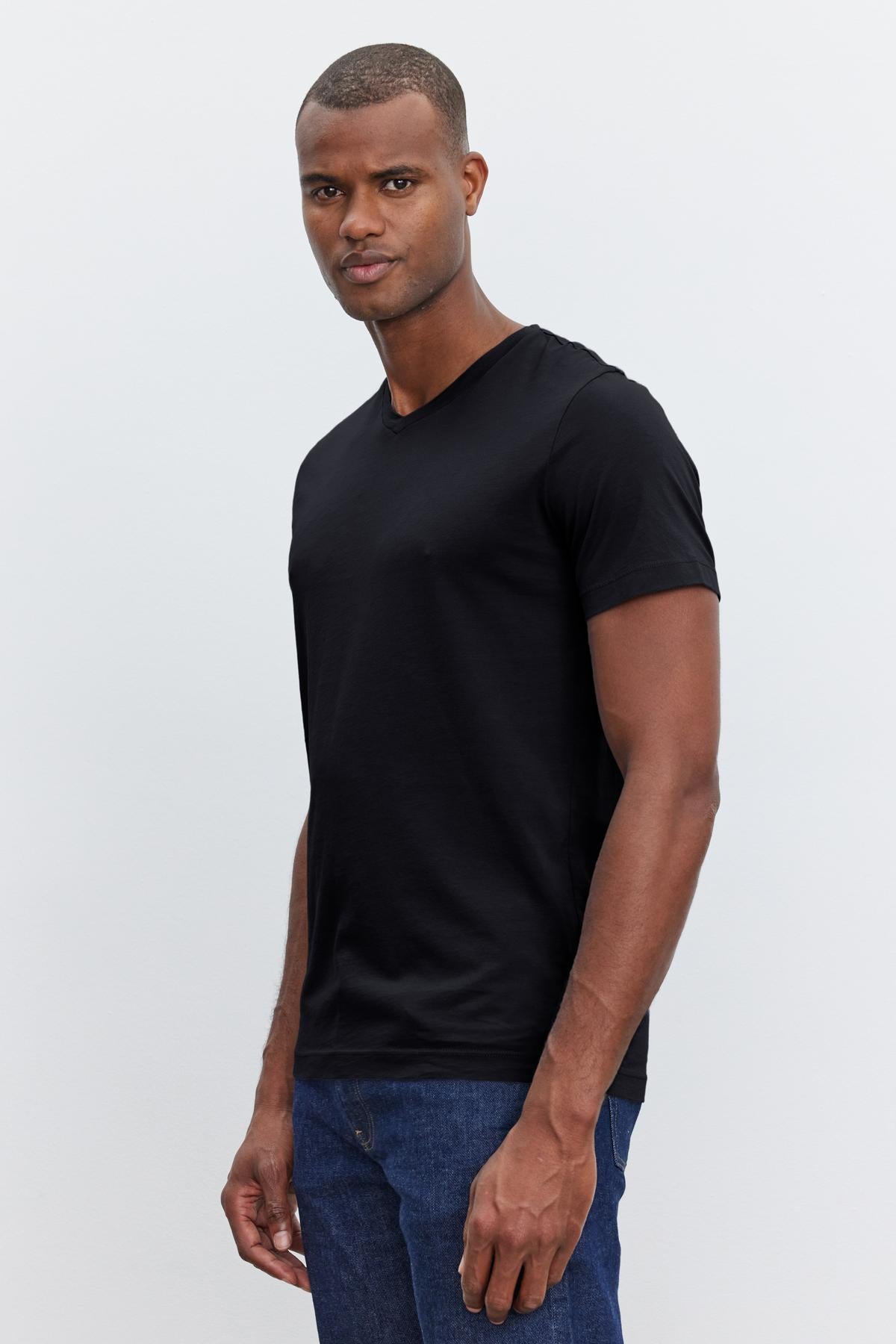   A man wearing a Velvet by Graham & Spencer SAMSEN TEE with a flawless fit and blue jeans stands in front of a plain light gray background. He is looking directly at the camera with a neutral expression. 