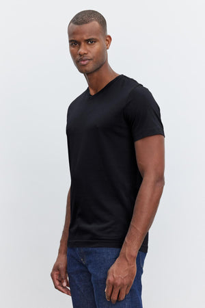 A man wearing a Velvet by Graham & Spencer SAMSEN TEE with a flawless fit and blue jeans stands in front of a plain light gray background. He is looking directly at the camera with a neutral expression.