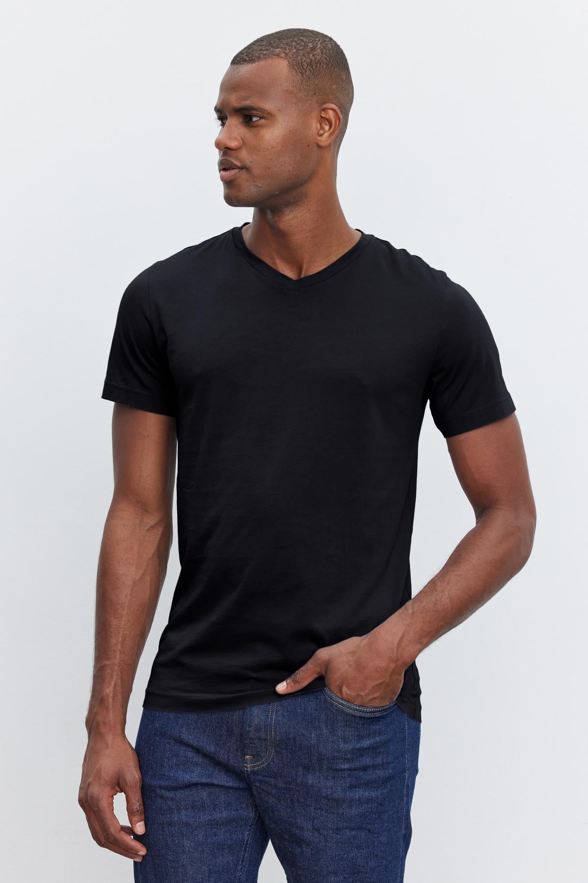   A man stands against a plain background, wearing a black SAMSEN TEE from Velvet by Graham & Spencer and blue jeans with a flawless fit. One hand rests in his pocket as he looks off to the side. 