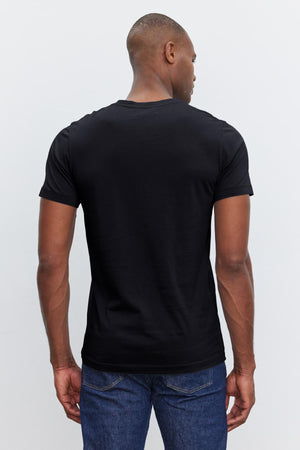 A person wearing a plain black SAMSEN TEE by Velvet by Graham & Spencer and blue jeans, showcasing a flawless fit, is shown from the back.