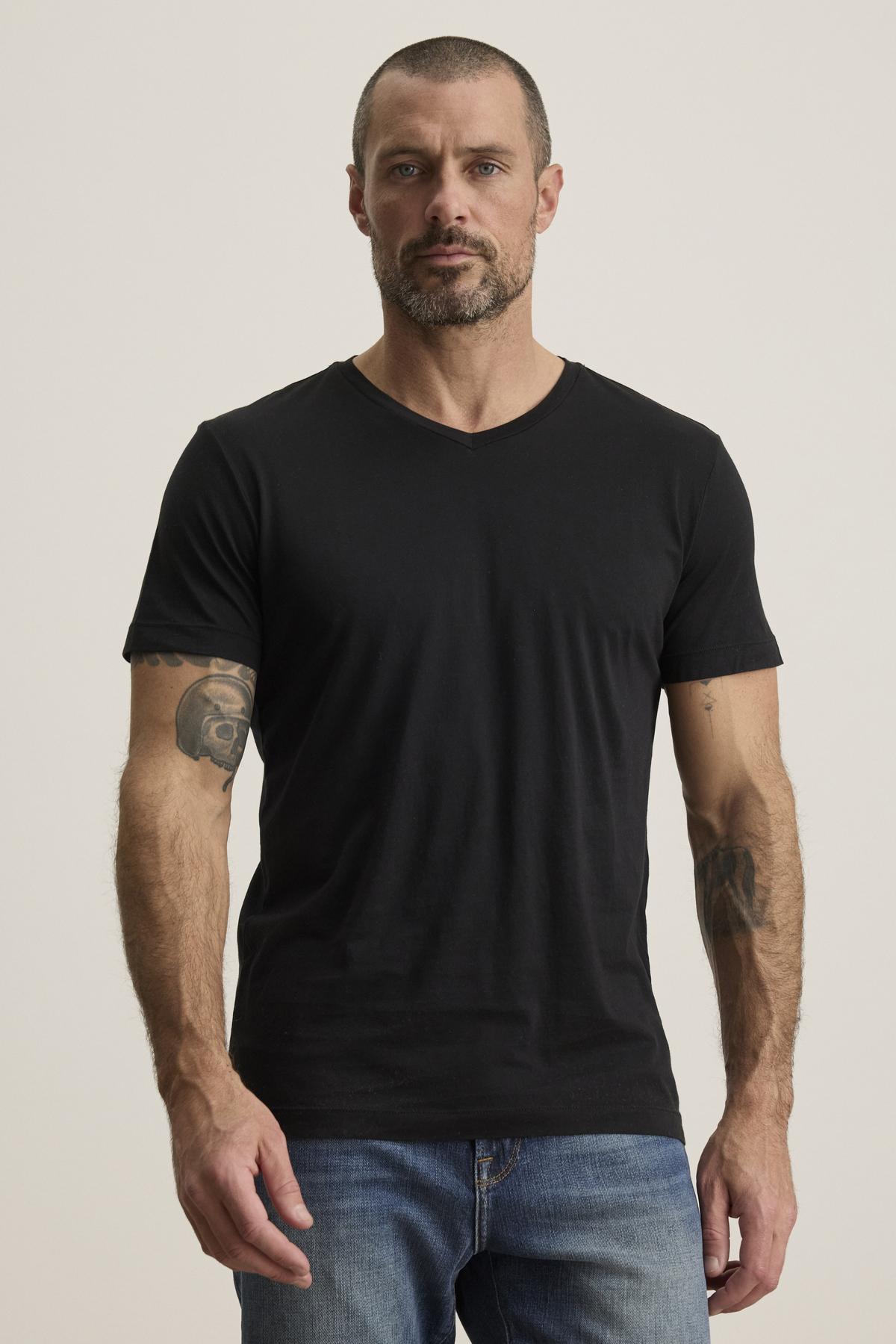 A man with short hair and tattoos on both arms is wearing a SAMSEN TEE in black by Velvet by Graham & Spencer, crafted from comfortable Pima jersey, along with jeans, as he stands against a plain background.-38504012579009