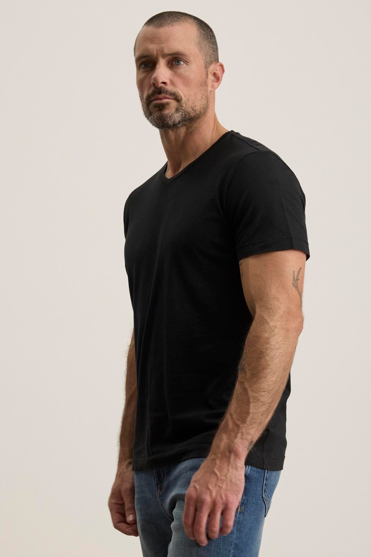   A man in a black SAMSEN TEE by Velvet by Graham & Spencer with a v-neckline and blue jeans stands against a plain background. 