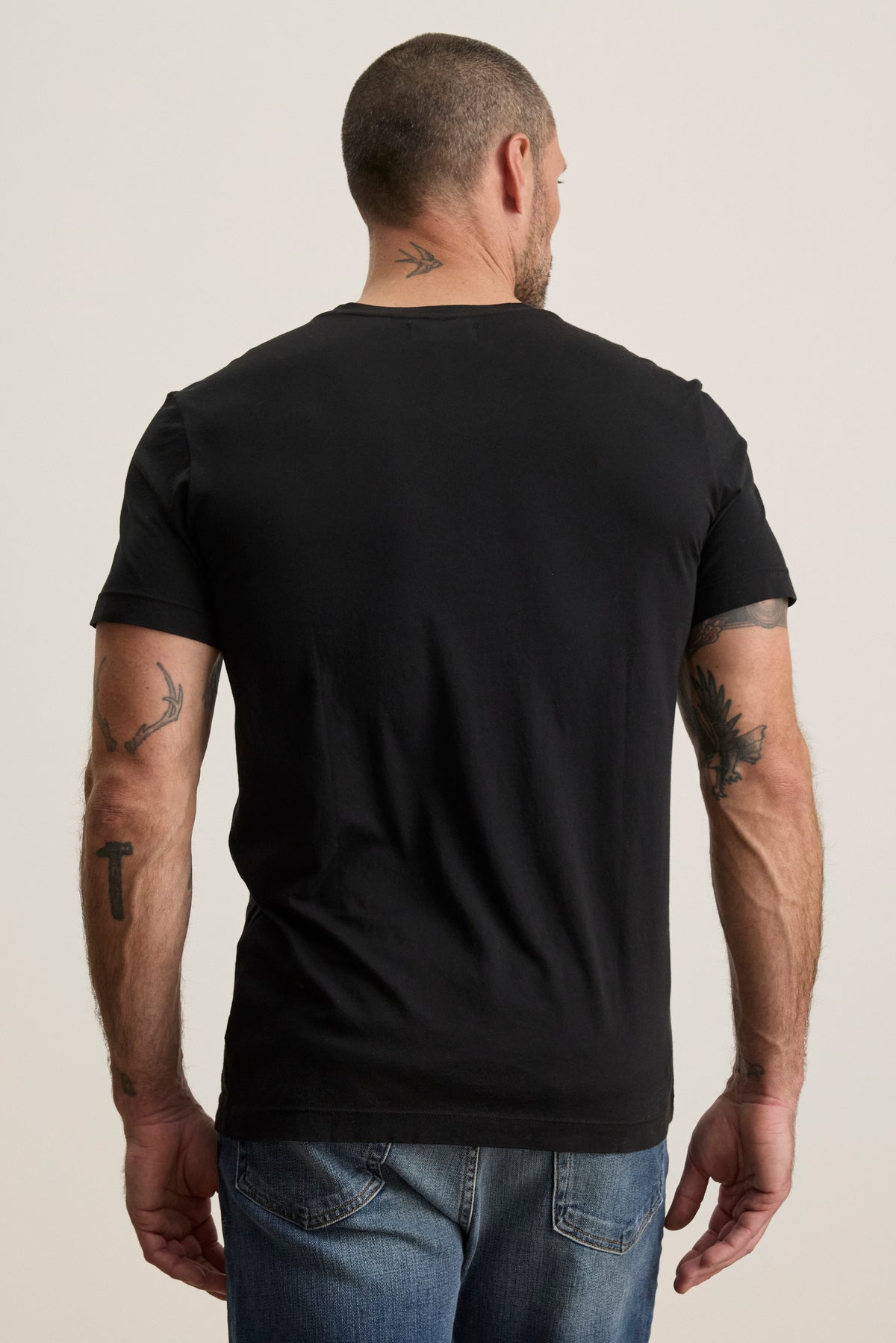   A man with short hair, facing away, wears the SAMSEN TEE by Velvet by Graham & Spencer—a black Pima jersey v-neck t-shirt paired with jeans—accentuating his arm and neck tattoos. 