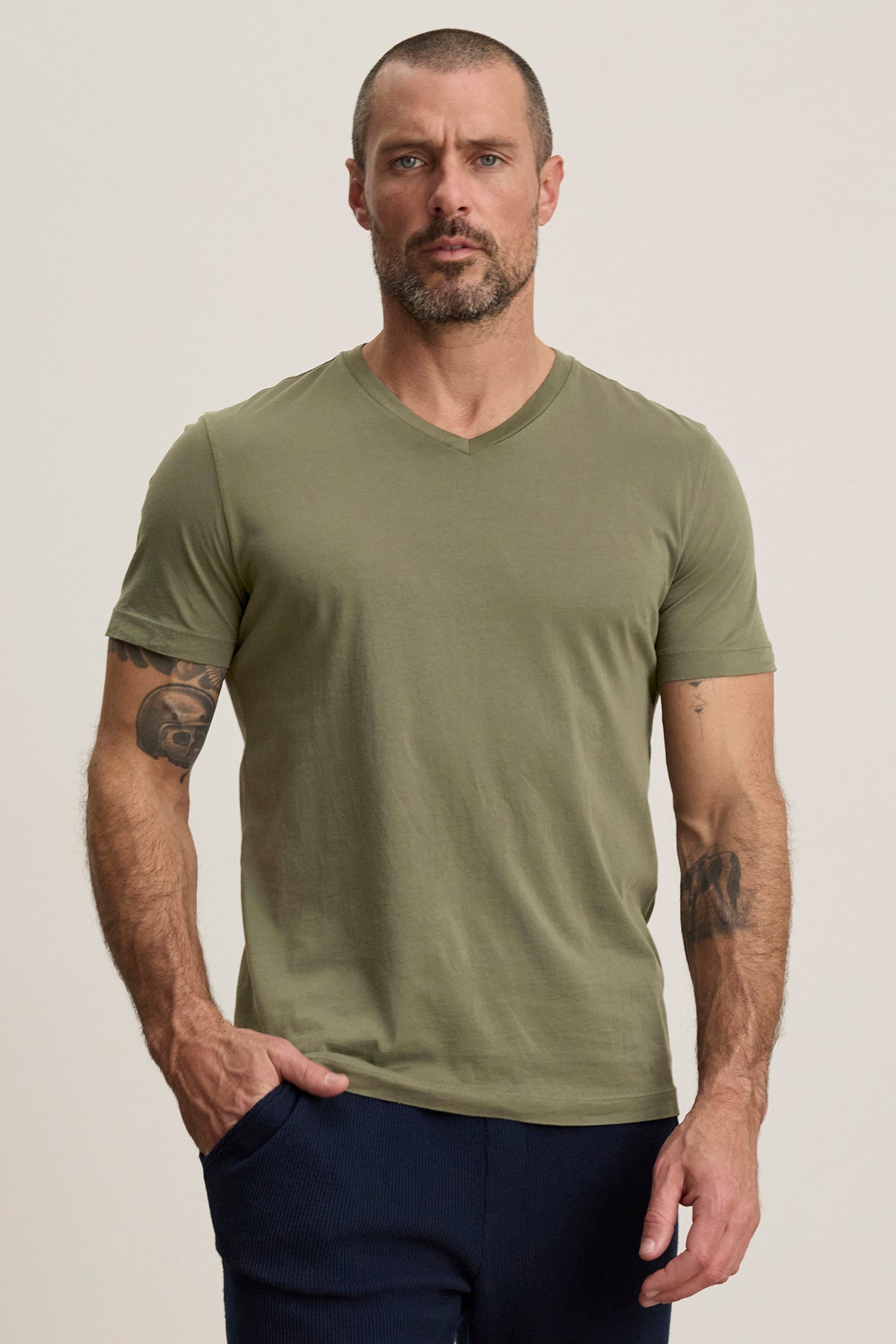   A bearded, tattooed man wears the Velvet by Graham & Spencer SAMSEN TEE, a green Pima jersey v-neck t-shirt, with dark pants. Standing against a plain background with one hand in his pocket, this tee is ideal for layering in cooler weather. 