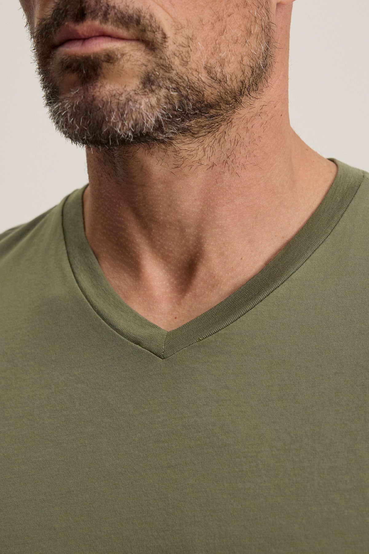   The SAMSEN TEE from Velvet by Graham & Spencer is showcased in a close-up of a man wearing the green Pima jersey V-neck T-shirt. His salt-and-pepper beard adds texture, complemented by a plain light gray background. 