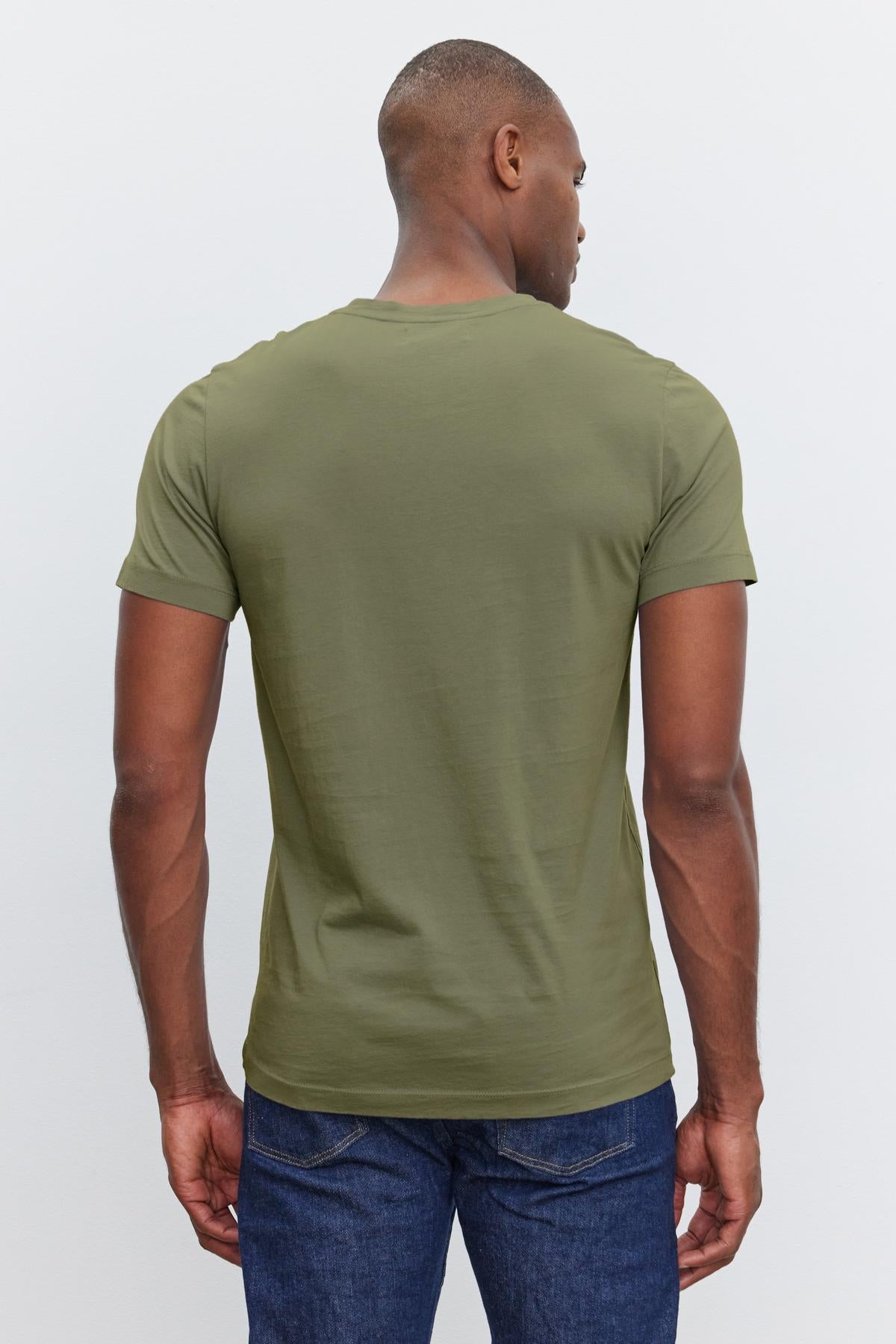   A person wearing the SAMSEN TEE from Velvet by Graham & Spencer, styled in green with a V-neckline, paired with blue jeans for a laid-back look, is standing with their back to the camera. 