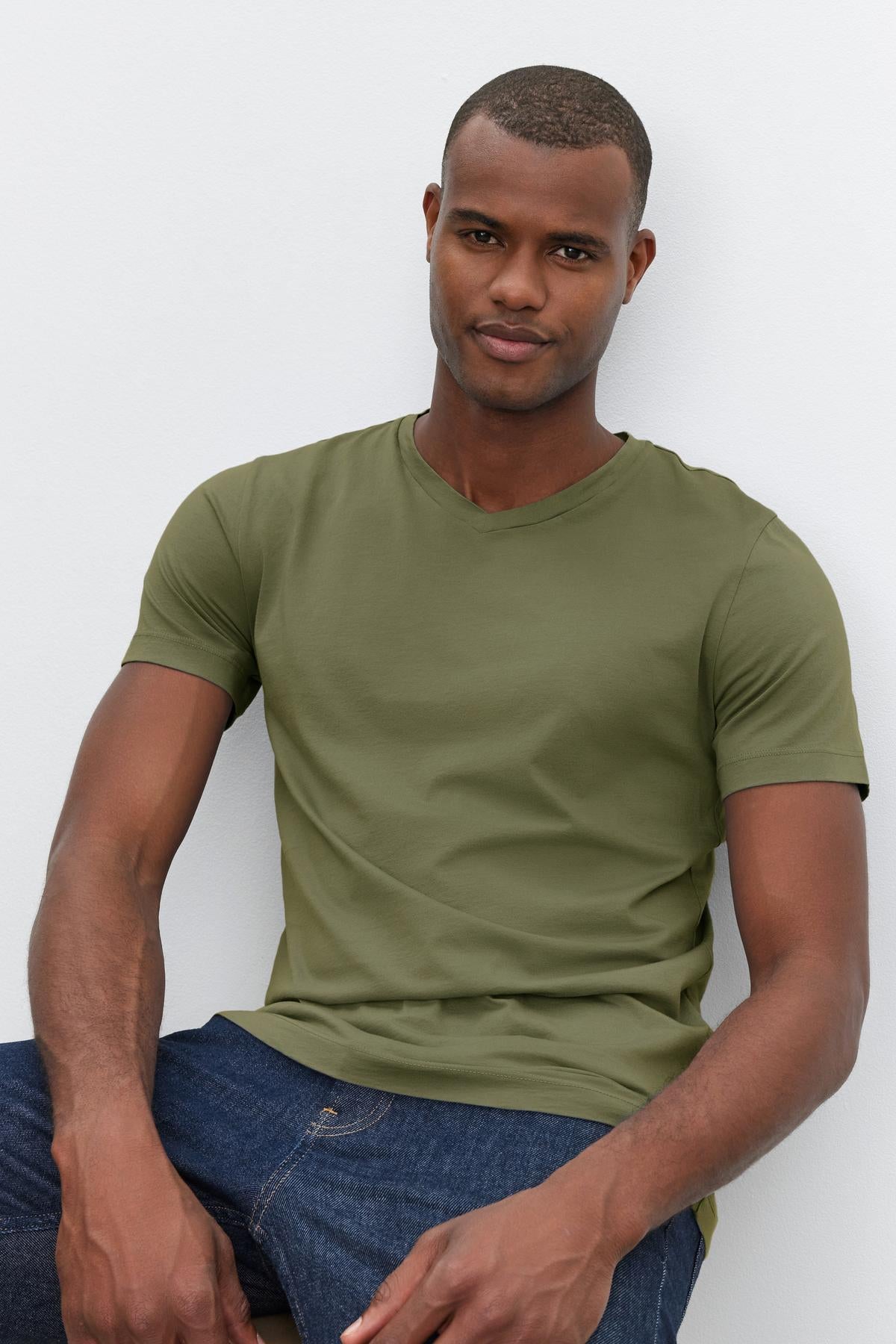 A person is wearing a green SAMSEN TEE by Velvet by Graham & Spencer and blue jeans, sitting against a light gray background.-38264837374145