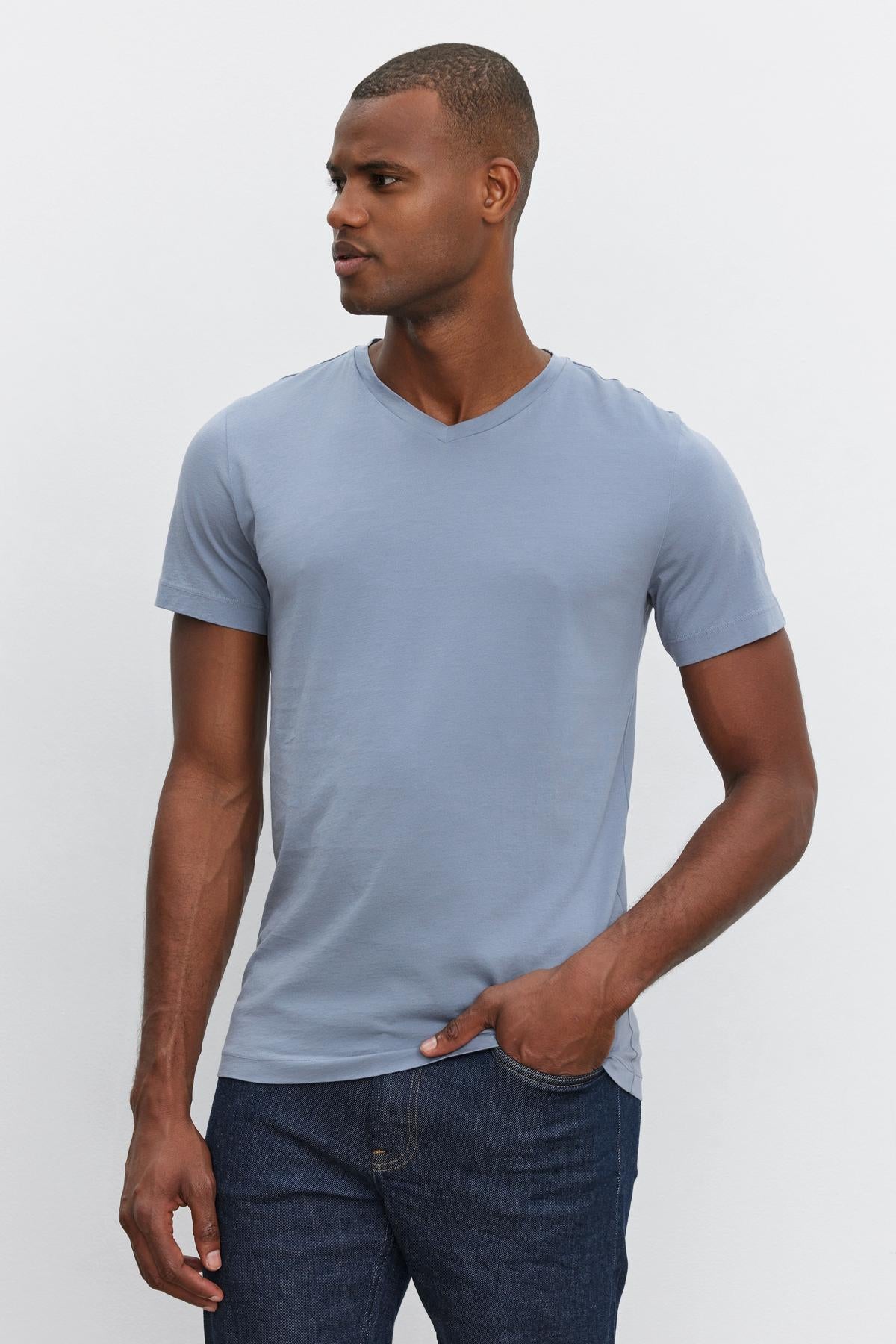   A man wearing a light blue SAMSEN TEE by Velvet by Graham & Spencer made from whisper cotton knit and dark blue jeans stands against a plain white background, looking to his left. 