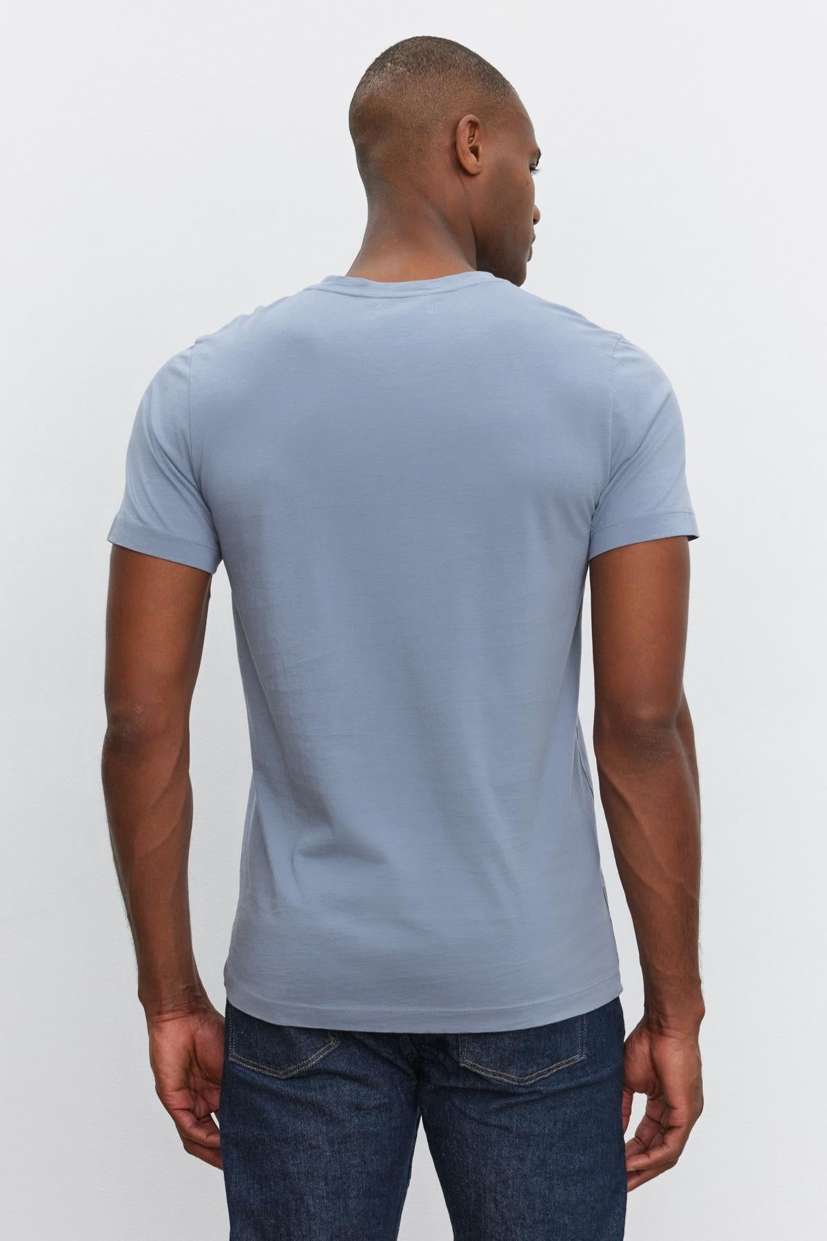   A man, clad in everyday wear— a light blue short-sleeve SAMSEN TEE made of whisper cotton knit by Velvet by Graham & Spencer and dark jeans, faces away from the camera. 