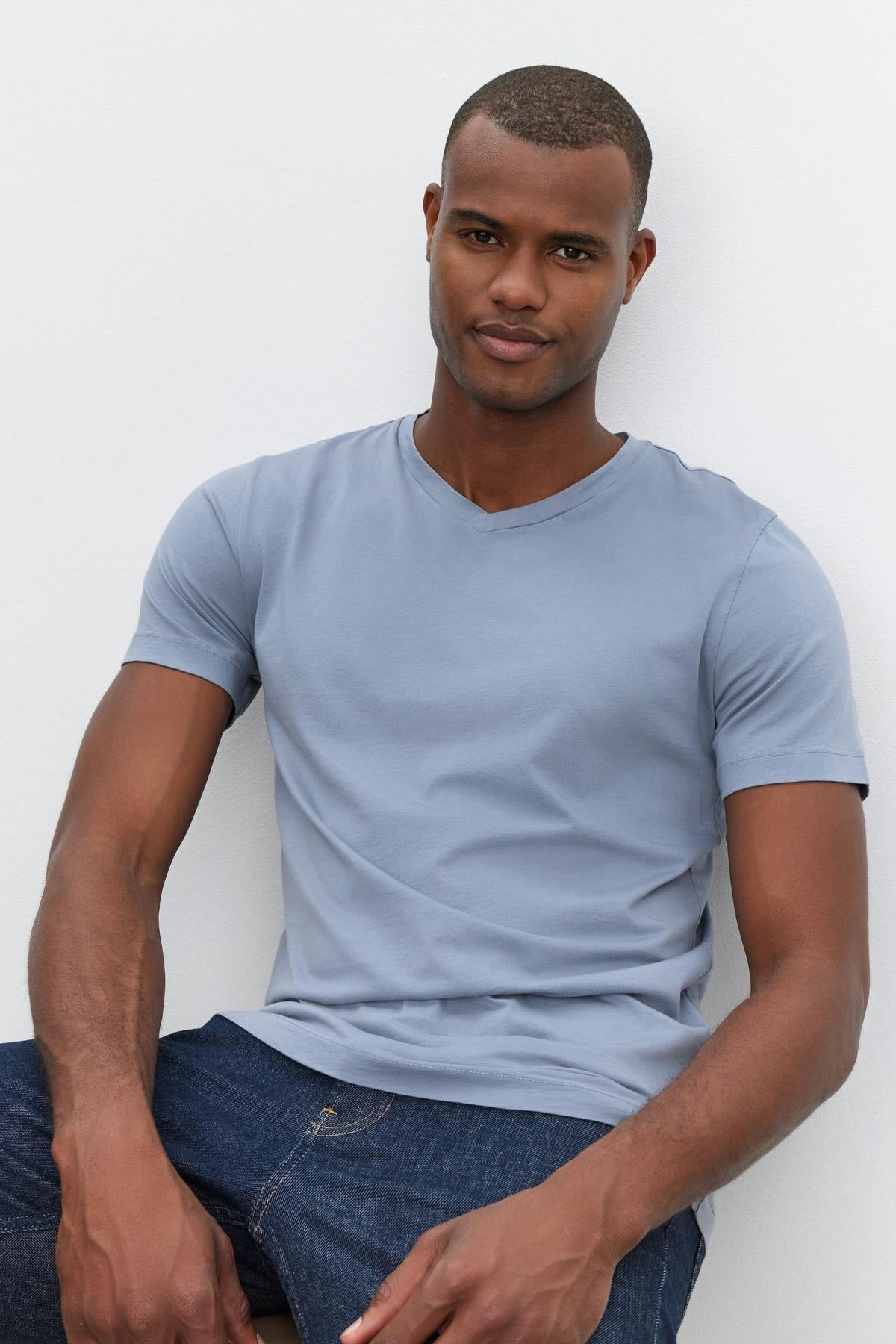   A man in a light blue SAMSEN TEE by Velvet by Graham & Spencer with a whisper cotton knit and dark jeans is seated against a plain white background. 