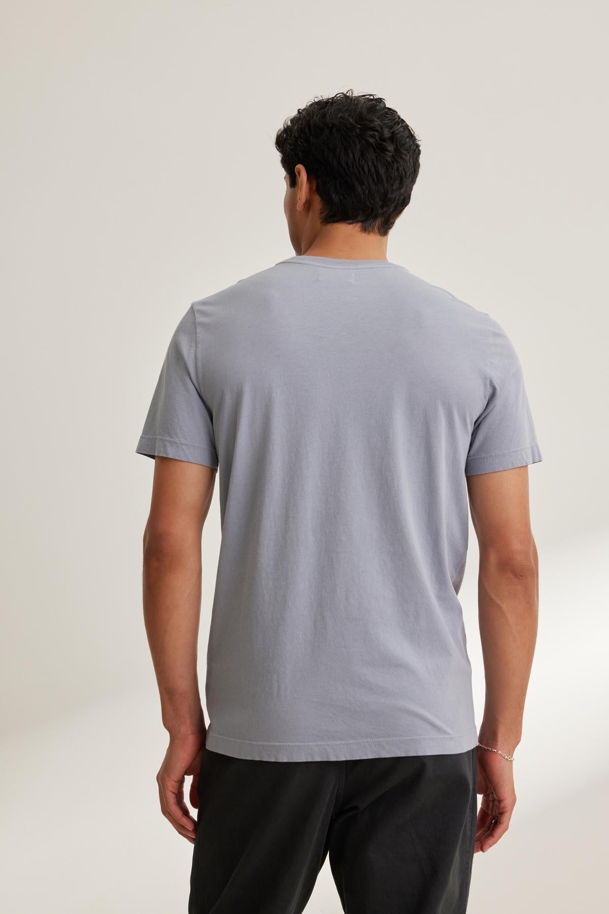   A person, with their back to the camera, wears a Velvet by Graham & Spencer SAMSEN TEE in light gray Pima jersey featuring a v-neckline, paired with dark pants against a plain background. 