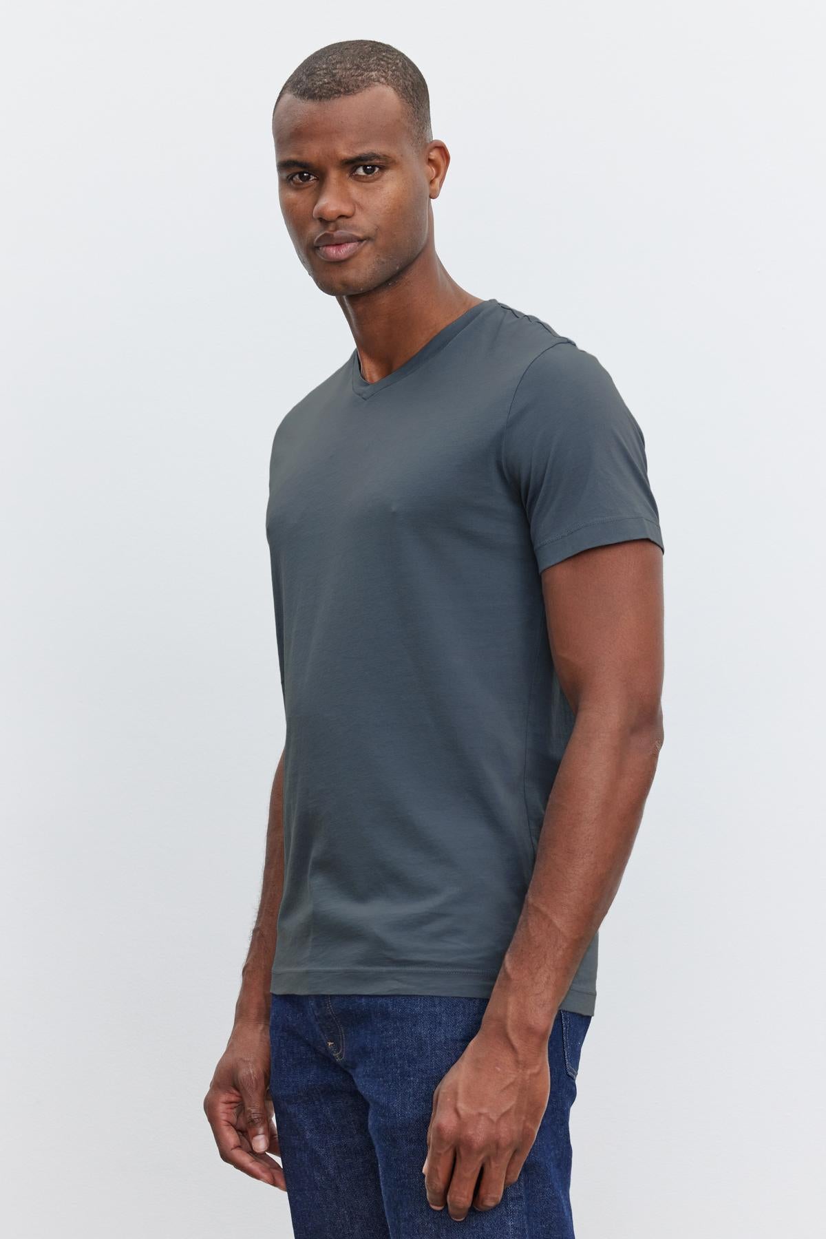   A man wearing a dark gray short-sleeve, v-neckline SAMSEN TEE by Velvet by Graham & Spencer and blue jeans stands against a plain light background. He is looking directly at the camera, embodying effortless everyday wear. 