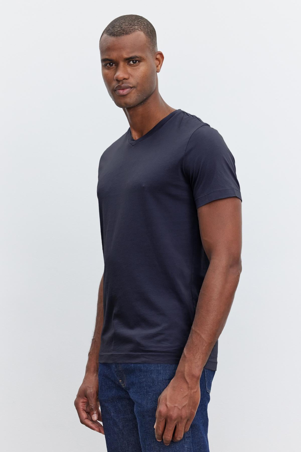   A man wearing a dark-colored Velvet by Graham & Spencer SAMSEN TEE and jeans stands against a plain white background. The V-neckline adds subtle detail as he looks directly at the camera with a neutral expression, showcasing a flawless fit. 