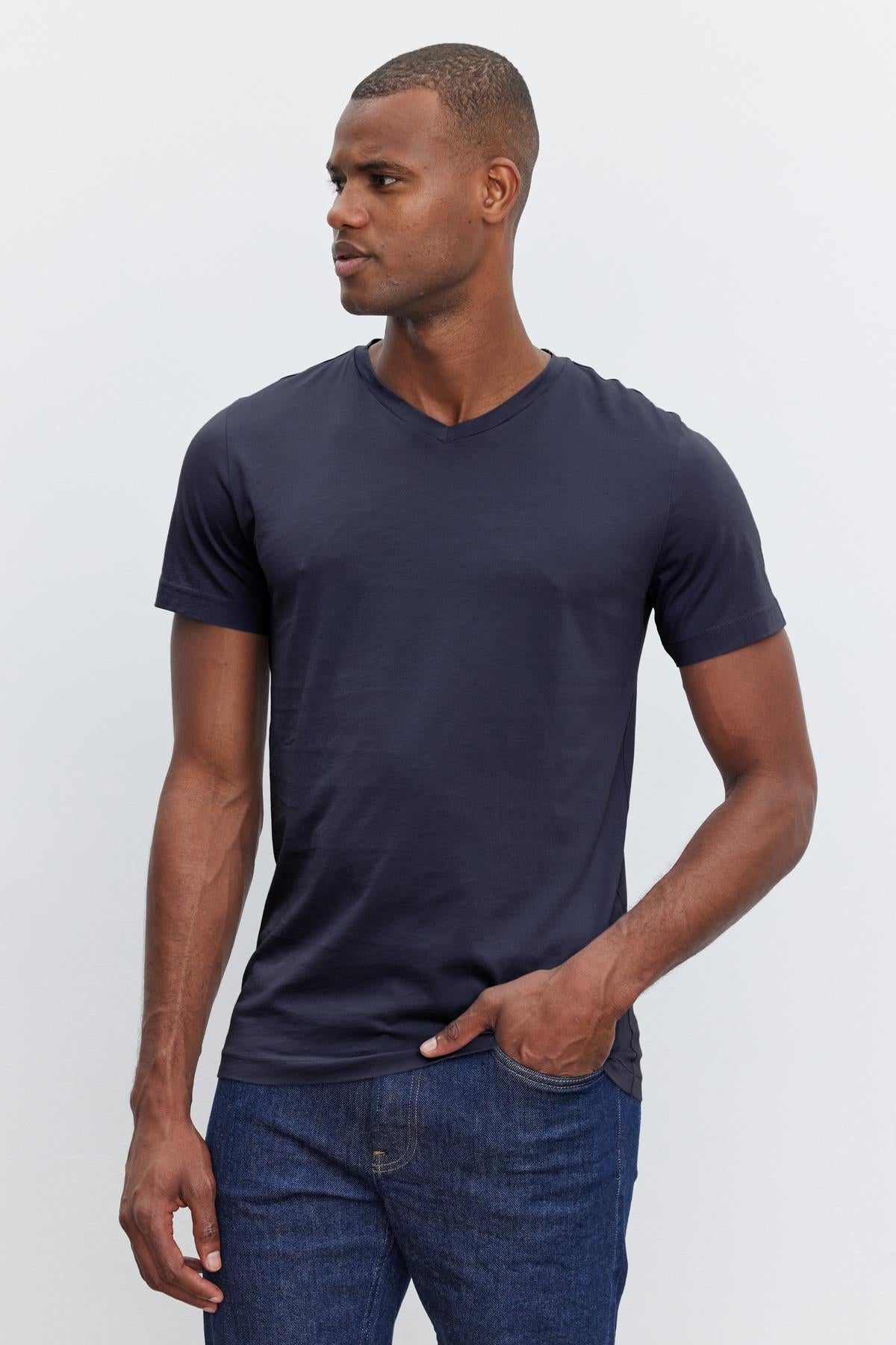   A man wearing a dark blue Velvet by Graham & Spencer SAMSEN TEE and blue jeans stands against a plain light background, with his right hand in his pocket. He is looking to his left. 