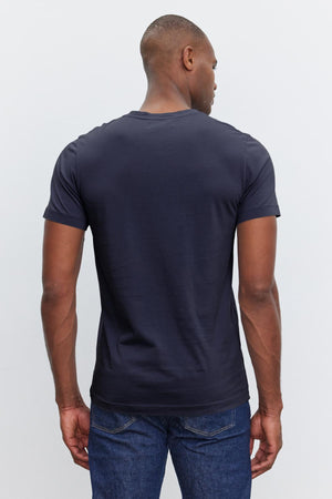 Man wearing a dark navy short-sleeve SAMSEN TEE by Velvet by Graham & Spencer with a whisper knit for a flawless fit and blue jeans, facing away from the camera, with his head turned slightly to the left.