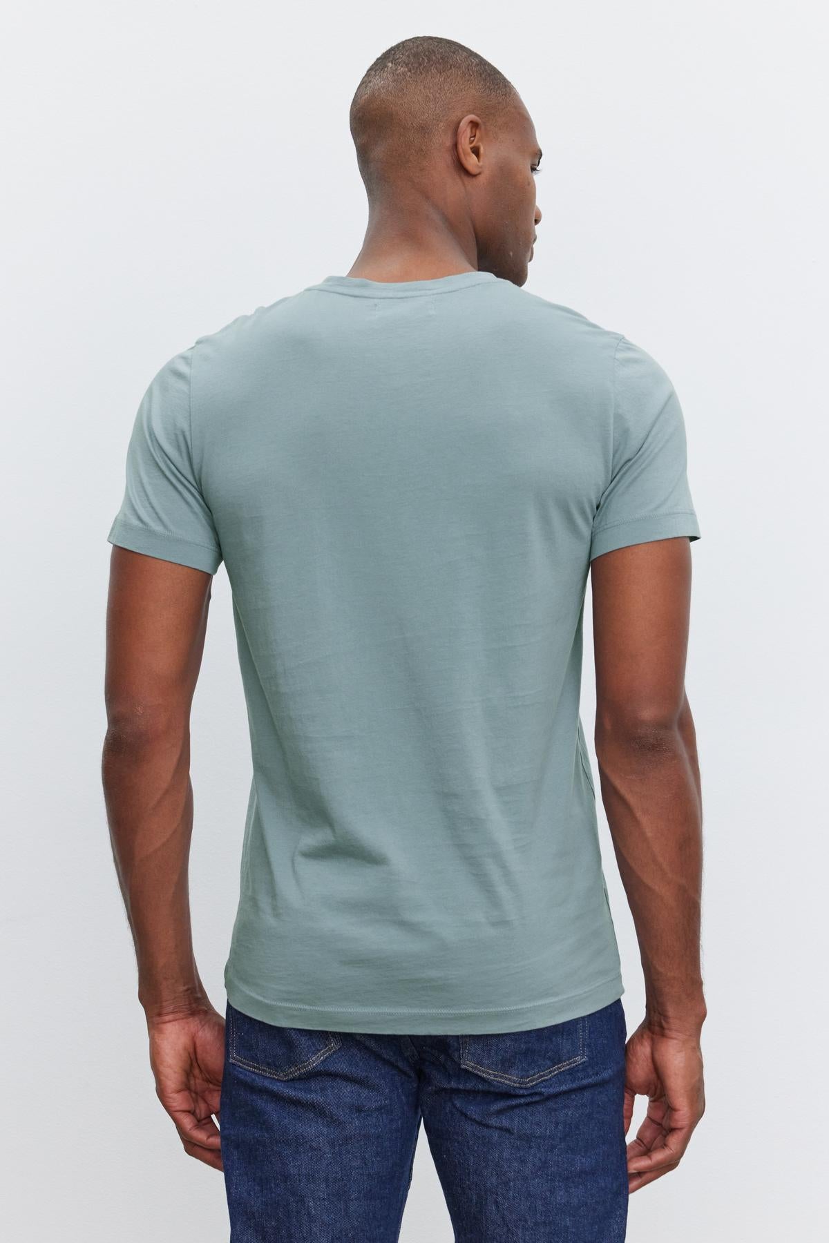 A person is standing with their back to the camera, wearing a light blue SAMSEN TEE by Velvet by Graham & Spencer and dark blue jeans against a plain white background.-37409982677185