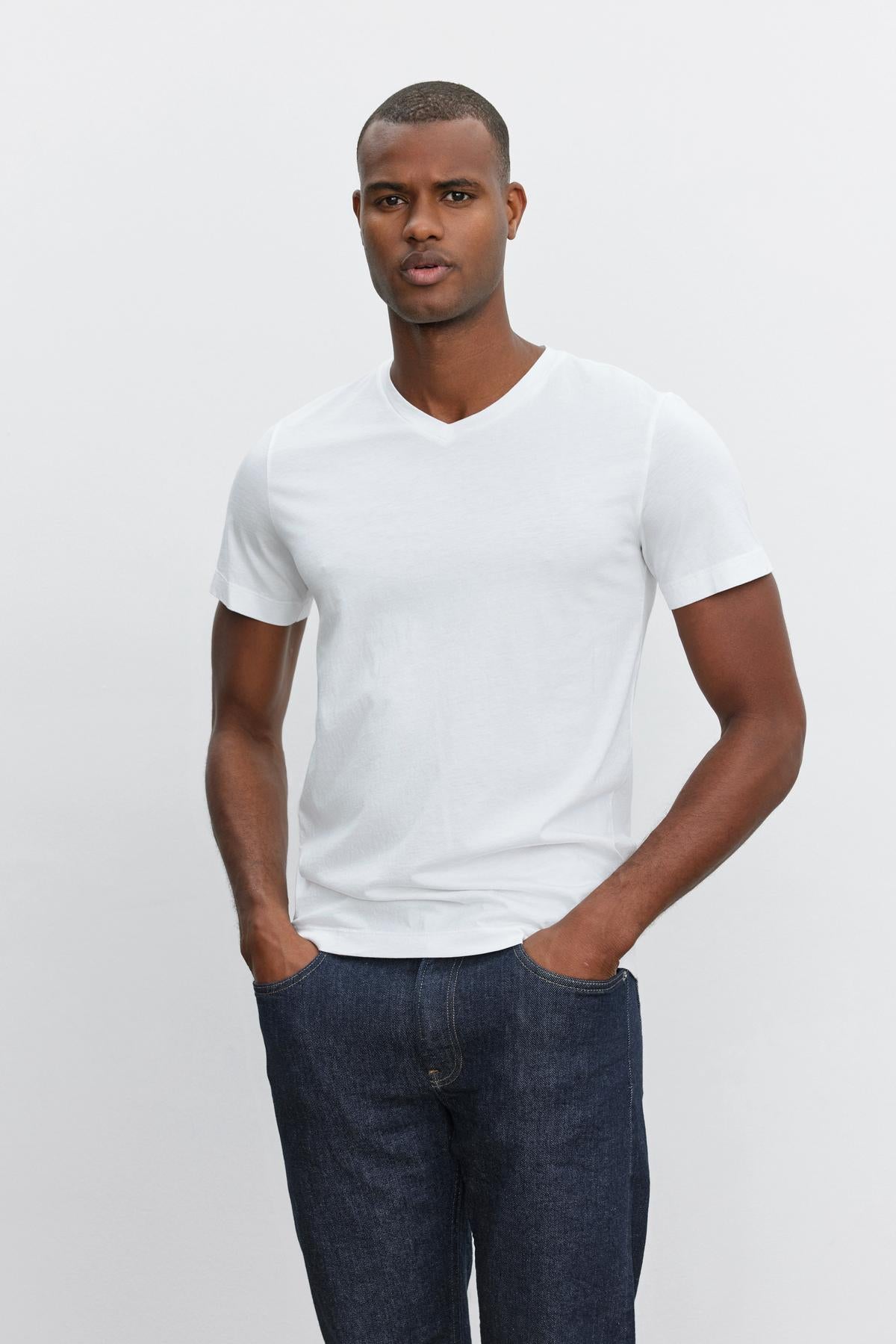   A man stands against a plain background, wearing a white SAMSEN TEE from Velvet by Graham & Spencer with a flawless fit and dark jeans. He has his hands in his pockets. 