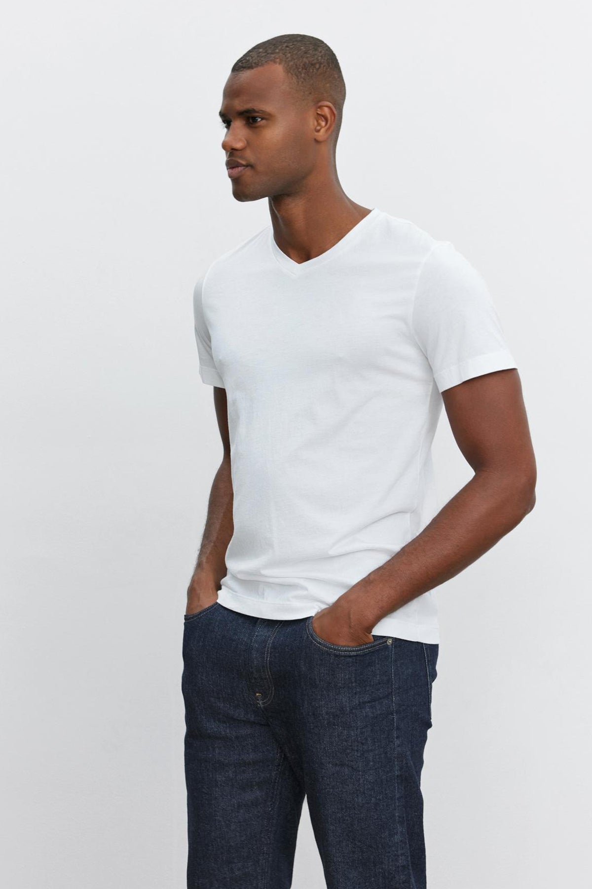 A man dressed in a white SAMSEN TEE by Velvet by Graham & Spencer and dark blue jeans, standing with his hands in his pockets against a plain light background.-35867446018241