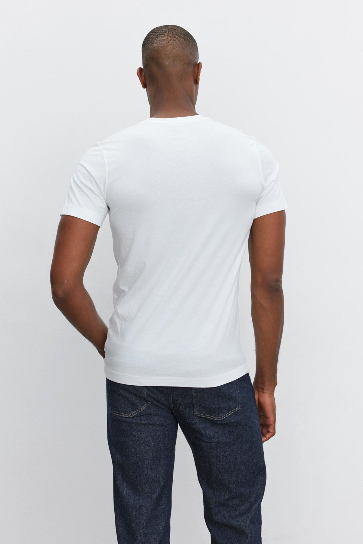   A man is standing with his back facing the camera, wearing a Velvet by Graham & Spencer SAMSEN TEE with a V-neckline and dark jeans against a white background, showcasing a flawless fit. 