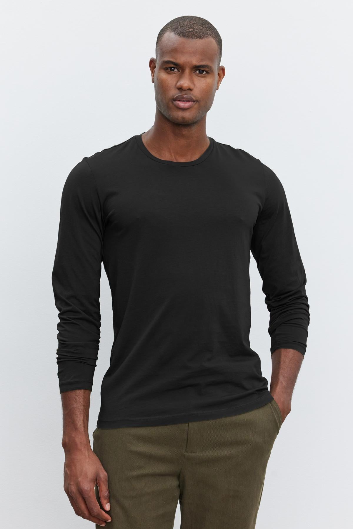   A man wearing a black long-sleeve SKEETER TEE by Velvet by Graham & Spencer and olive pants stands against a plain white background with one hand in his pocket, showcasing a versatile wardrobe addition. 