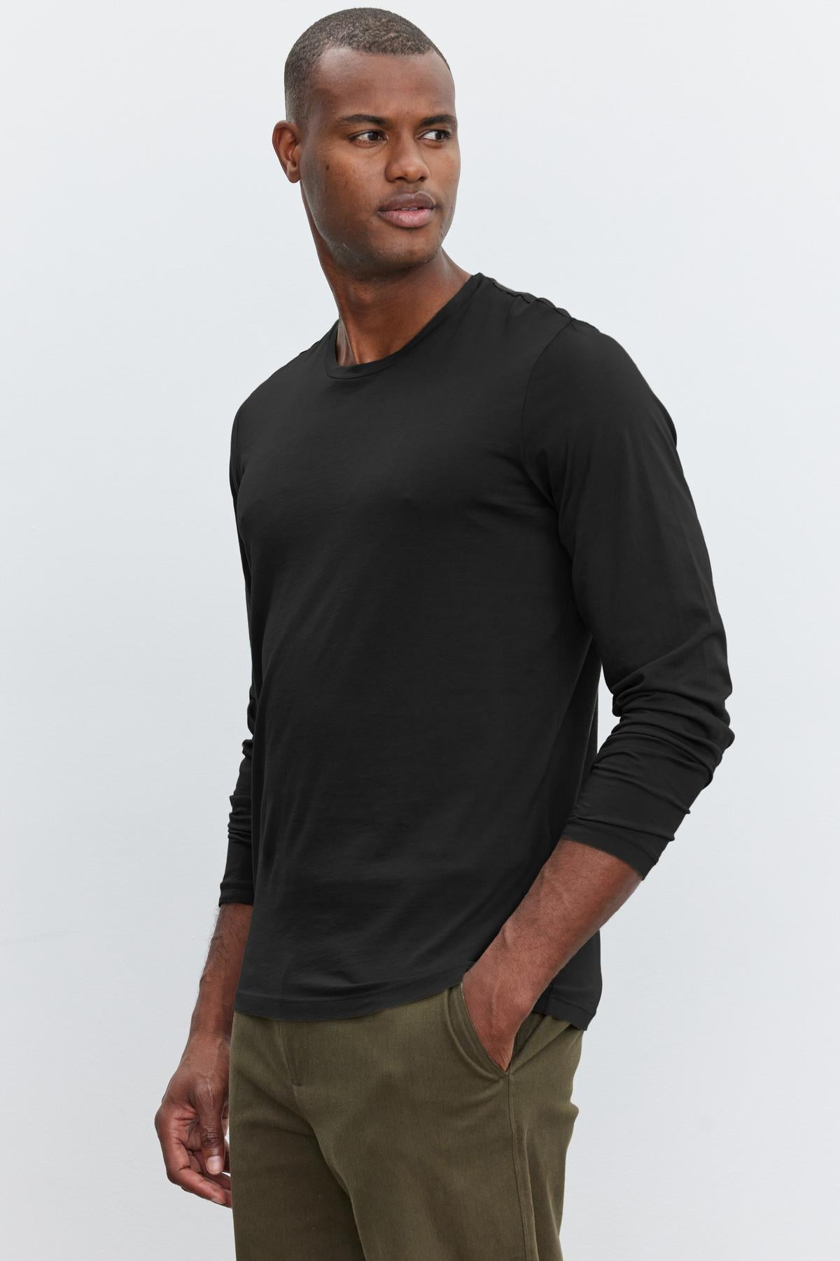   A person wearing a black long-sleeved SKEETER TEE by Velvet by Graham & Spencer and olive-green pants stands with one hand in their pocket, looking to the side. This versatile wardrobe addition, made from whisper cotton knit, complements their relaxed posture perfectly. 