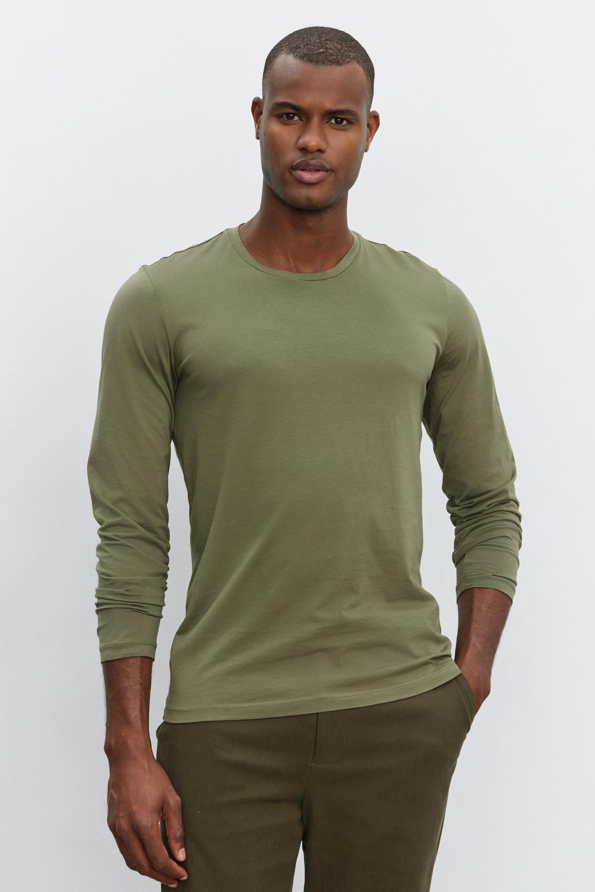 A person standing against a plain backdrop, wearing a long-sleeve, olive green SKEETER TEE by Velvet by Graham & Spencer made of Pima cotton and matching pants, with a neutral expression and hands in pockets.-37682145689793