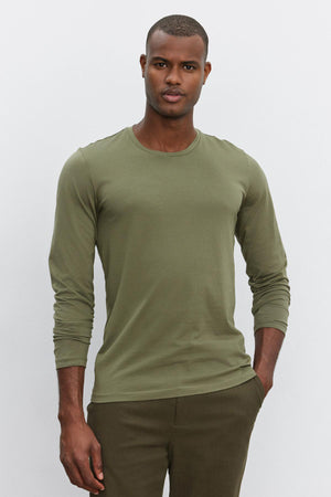 A person standing against a plain backdrop, wearing a long-sleeve, olive green SKEETER TEE by Velvet by Graham & Spencer made of Pima cotton and matching pants, with a neutral expression and hands in pockets.