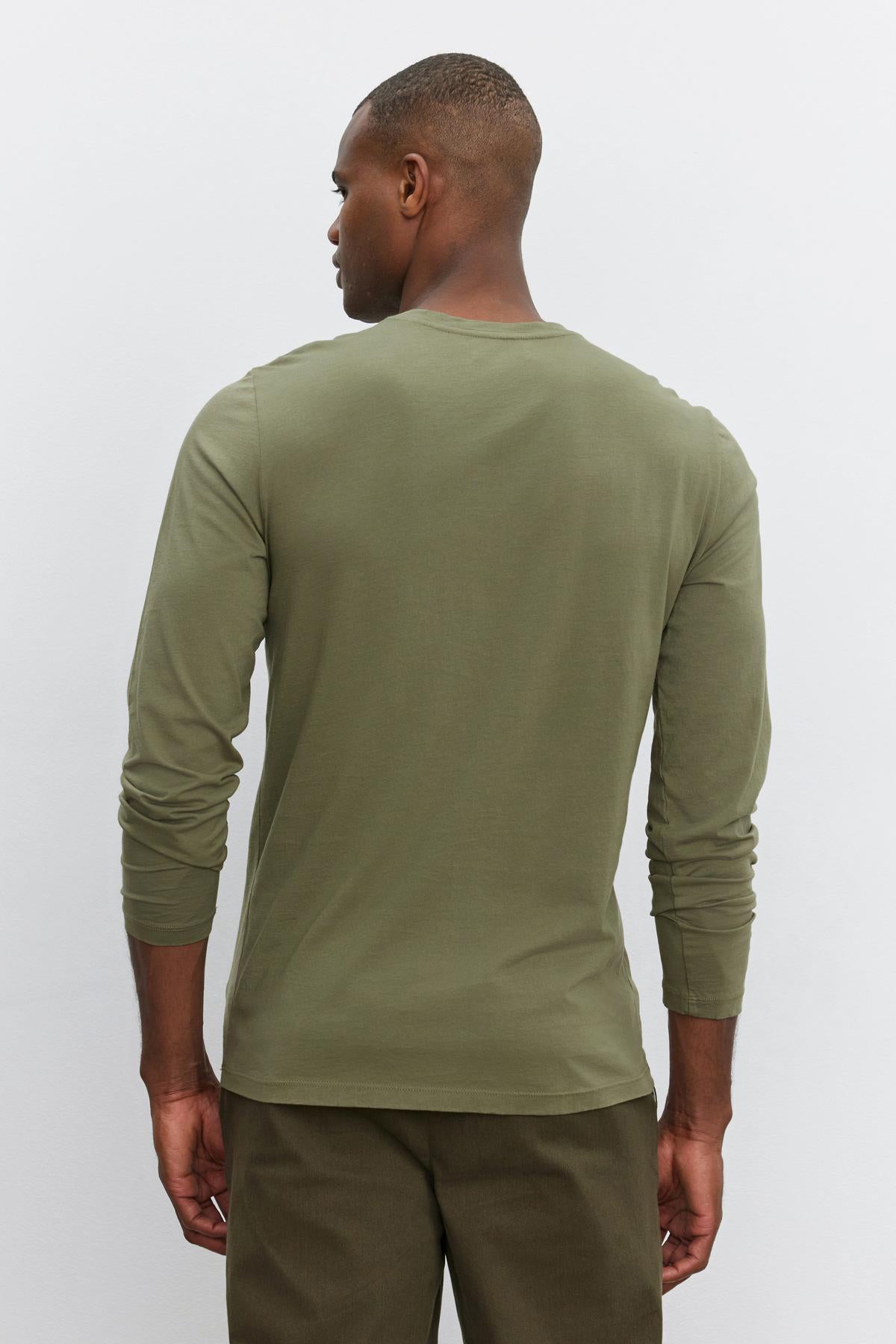   A man stands facing away from the camera, wearing a long-sleeved olive green SKEETER TEE by Velvet by Graham & Spencer with a classic crew neckline and matching pants against a plain light background. 