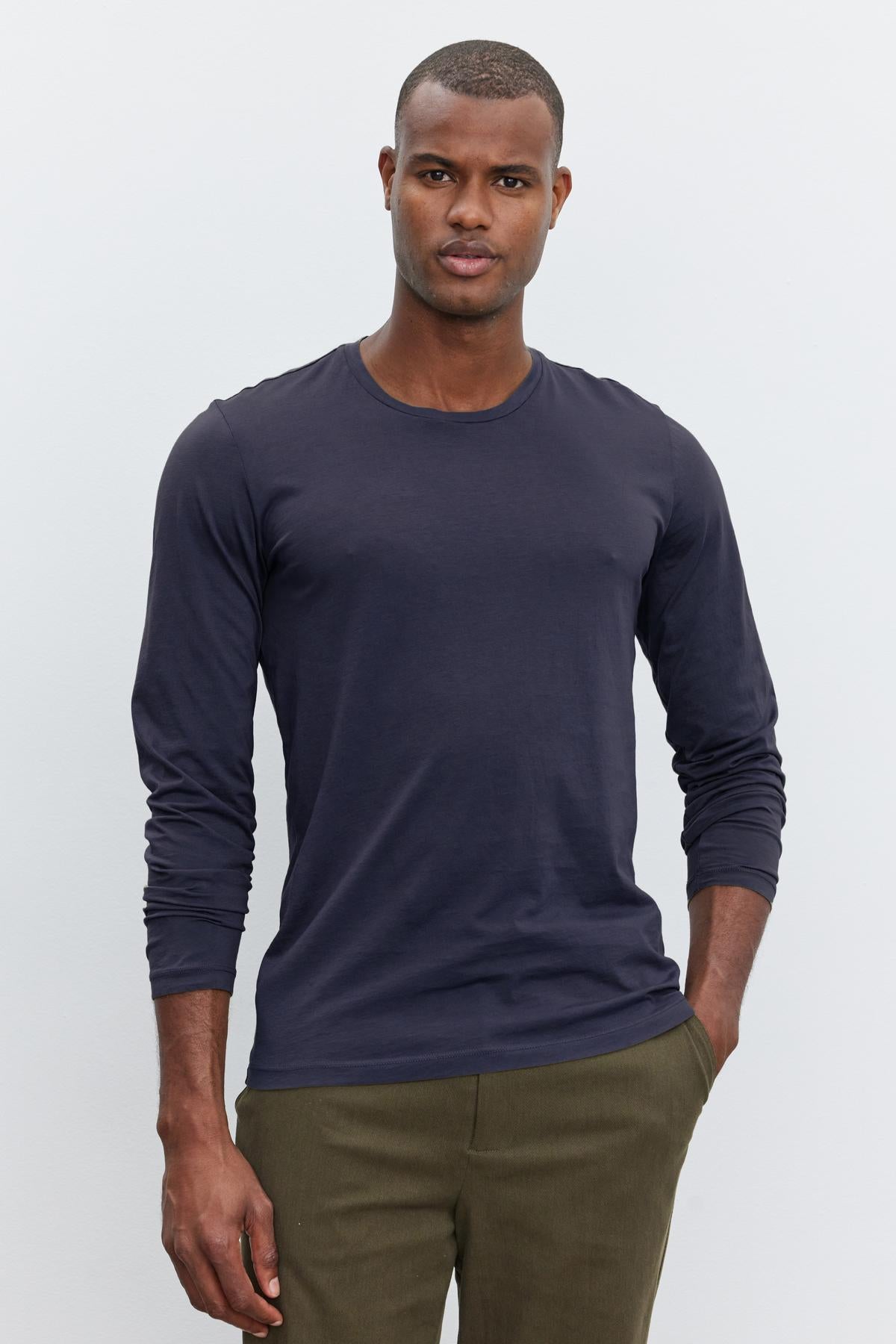   A person stands against a plain background, wearing a long-sleeved, navy blue SKEETER TEE from Velvet by Graham & Spencer and olive green pants, with hands in their pockets. 