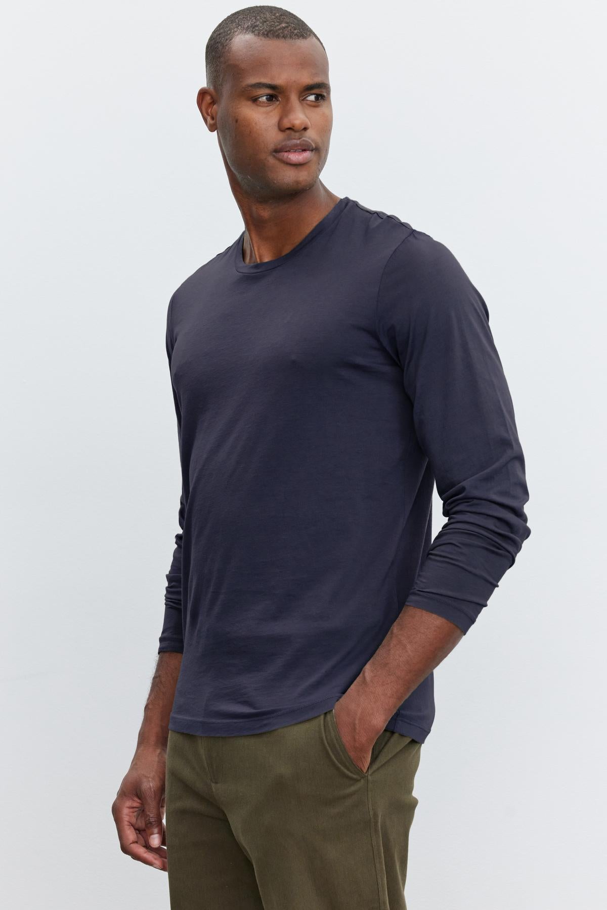   A man wearing a long-sleeved navy blue SKEETER TEE by Velvet by Graham & Spencer and olive green pants stands with one hand in his pocket, looking to the side against a plain background. His choice of attire is a versatile wardrobe addition, effortlessly blending comfort and style. 